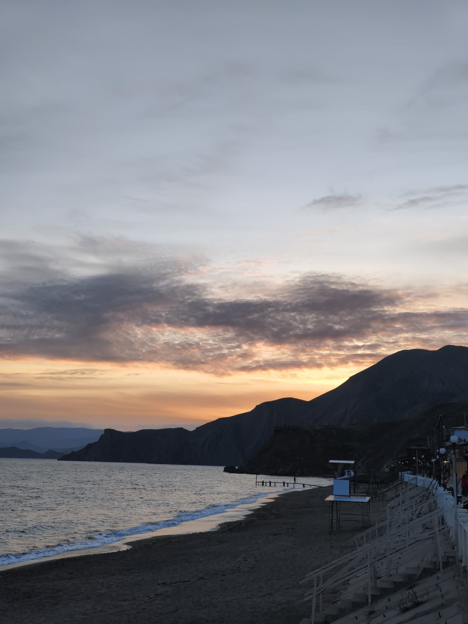 A scenic view of a coastline during sunset or twilight in the Орджоникидзевский поселковый совет, Ukraine. The foreground consists of a beach with dark sand, leading to a calm body of water, possibly a sea or bay. The water's edge is marked by small, gentle waves. Further back, there are mountains and coastal cliffs silhouetted against the colorful sky. A few structures, including a lifeguard stand, are visible on the beach or along the shore. There are also some wooden walkways or piers extending into the water. The sky exhibits a mix of colors, with orange, pink, and gray hues suggesting either a sunset or sunrise. Clouds are scattered across the sky, some illuminated by the sun's rays.