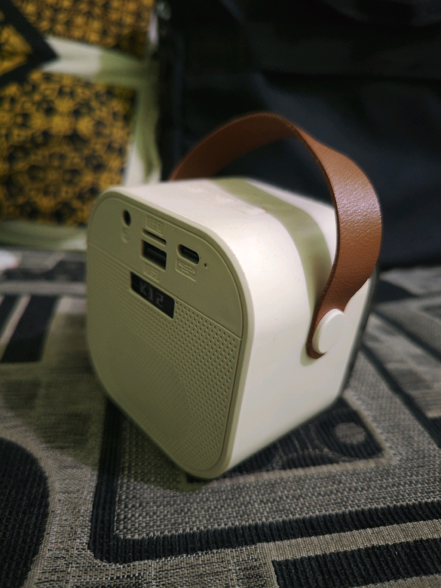 A beige-colored portable speaker with a brown leather handle is placed on a patterned surface. The speaker has a mesh front where the sound emanates from. On the top surface of the speaker, there are several ports, including a USB port, an SD card slot, and a charging port. A small digital display on the speaker shows "K12". The background is out of focus and appears to be a dark cloth, possibly a bag, and a yellow and black patterned fabric. The scene is indoors, and the lighting suggests it is daytime. There are no people visible in the image. The location of Sroyo, Indonesia is not visually represented in this image.