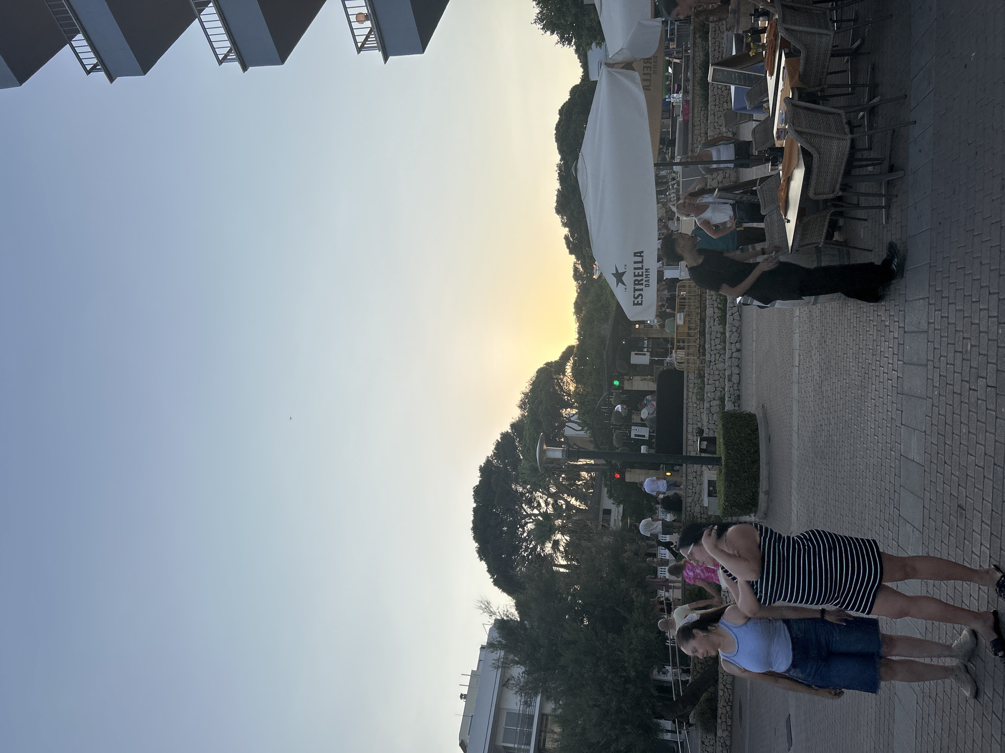 The image depicts an outdoor scene in Cala Rajada, Spain, during what appears to be sunset. In the foreground, two women stand facing towards the left. The woman on the left wears a blue tank top and denim shorts, while the woman on the right wears a black and white striped dress. Behind them, several tables and chairs are set up, some covered by large white umbrellas bearing the "ESTRELLA DAMM" logo. A man in a black t-shirt and pants is standing near one of the tables.
In the mid-ground, a group of people are gathered around a raised platform, possibly a stage, where some lighting equipment is visible. Trees and some buildings form the backdrop. The sky is clear and light blue, with a soft orange glow indicating the setting sun visible behind the trees. The ground is paved with rectangular stones. In the far right of the image, a portion of a multi-story building with balconies is visible against the sky.
