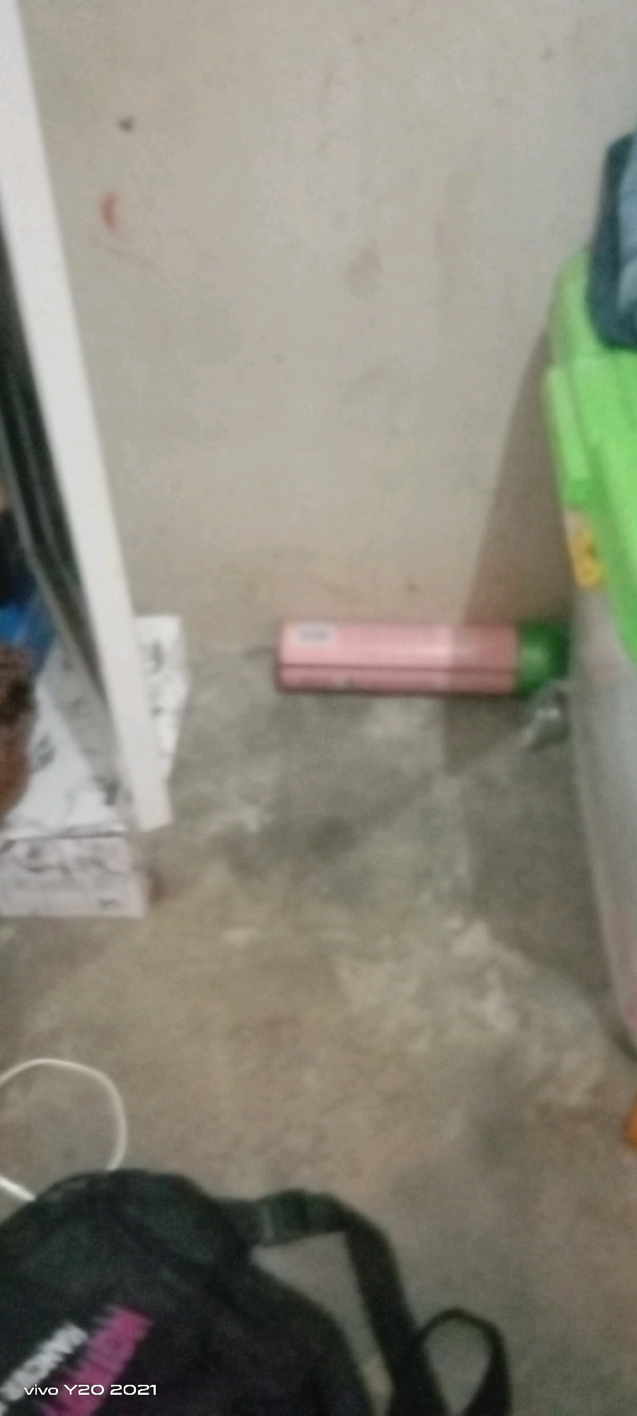 The image depicts a blurred indoor scene. On the right side, a portion of a light green object is visible, possibly a storage container, with a dark blue item resting on top. To the left of this, a pink rectangular object with a green end is lying horizontally on the floor. In the foreground on the left, a black backpack with white and pink lettering is partially visible. The floor appears to be concrete, with some discoloration and debris. A white vertical object, possibly a wall or partition, is visible on the far left. The overall impression is of a cluttered, utilitarian space, likely a room or storage area. No people or specific actions are discernible due to the blurriness of the image. The timestamp "vivo Y20 2021" suggests the photograph was taken in 2021 using a Vivo Y20 device.