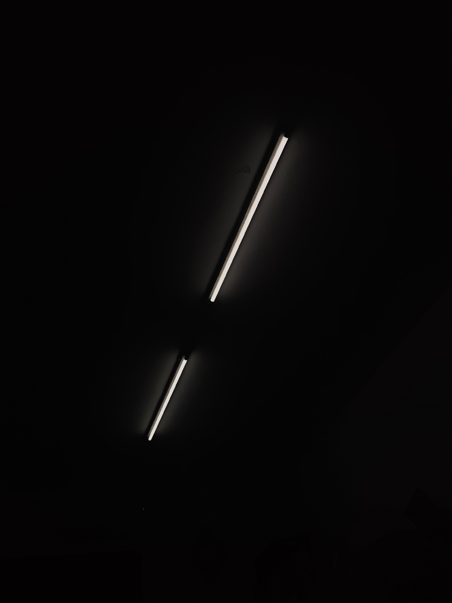 The image shows two fluorescent tube lights illuminated against a dark, nearly black background. The longer light is positioned diagonally in the upper right of the frame, while a shorter one is situated diagonally lower left. The environment appears to be indoors, and details are scarce due to the overall darkness. There are no visible people, subjects, actions, or interactions. The location of Chongqing High-tech Zone, China, is not evident from the image alone.