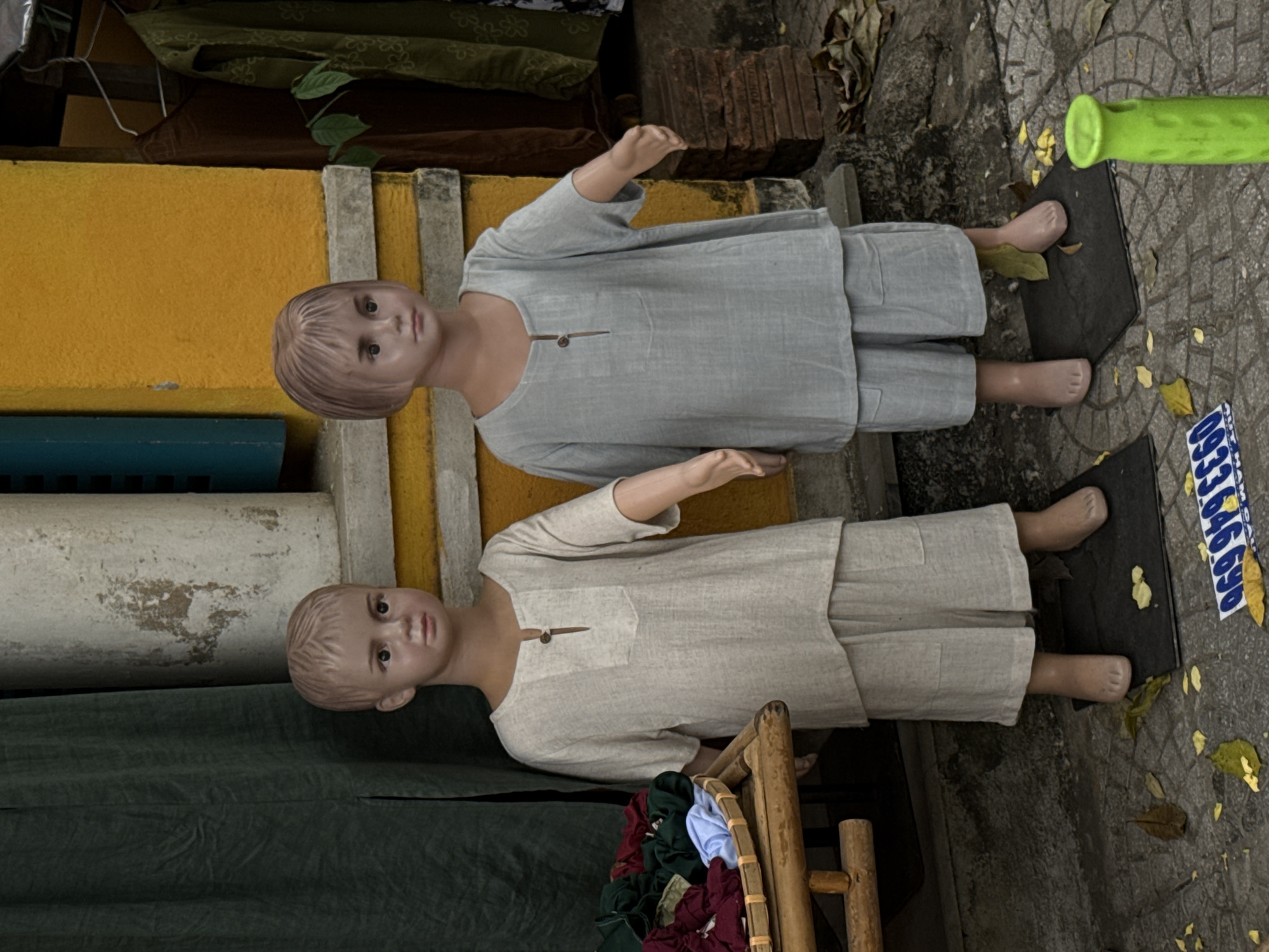 Two child-like mannequins are displayed upright on a sidewalk, likely outside a shop in Hội An Ward, Vietnam. The upper mannequin wears a light grey, loose-fitting tunic with matching pants, while the lower one is dressed in a similar style but in an off-white or beige color. Both tunics feature a small wooden toggle or button at the neckline. The mannequins have molded light brown hair and simple, painted facial features. Their feet are molded to resemble bare feet or skin-toned socks, and they stand on flat black bases.
The background on the left features a textured yellow wall, with a dark green fabric or tarp draped above the lower mannequin. A light-colored, weathered pipe or column is situated behind the lower mannequin's head. In the upper right, some stacked red bricks are visible. A small, bright green cylindrical object is partially seen in the upper right. To the right of the lower mannequin, a bamboo rack holds folded clothes in various colors, including red, blue, and green. The ground is a patterned sidewalk, scattered with dry, fallen leaves, suggesting an outdoor, daytime setting. A partially visible blue and white sign or sticker on the ground shows fragments of numbers, including "0933" and "6696", likely part of a phone number. The scene portrays a static street display for clothing.