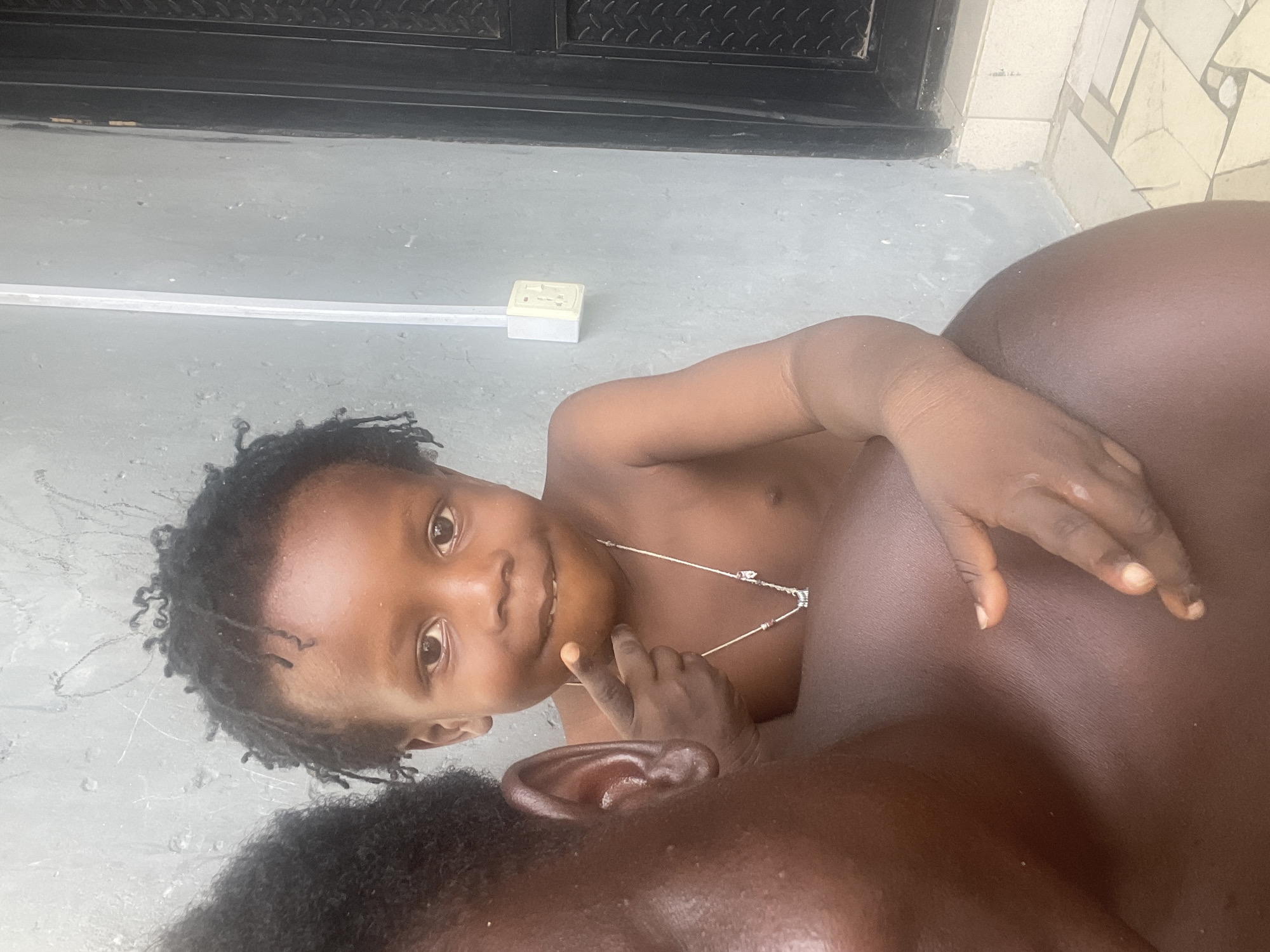 A child with dark skin and short, curly black hair is lying down, facing the camera and smiling. The child is wearing a delicate silver necklace. The child is lying on a dark-skinned adult, whose face is partially visible in the lower part of the frame. The adult has short black hair and is lying down. The background consists of a gray concrete floor and a black metal door. An electrical outlet is visible on the floor. The location is Badagry, Nigeria.