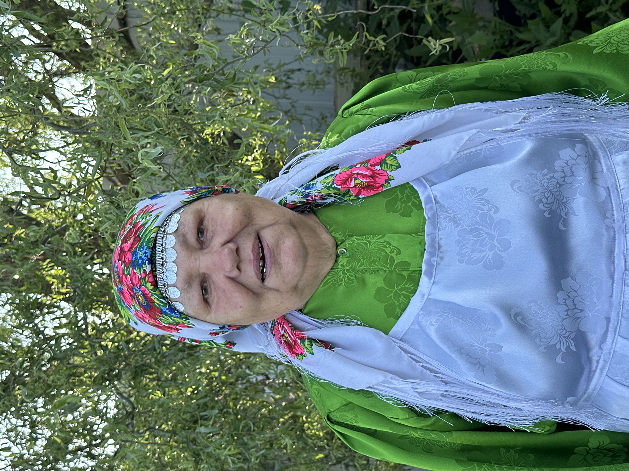 A medium shot, outdoor portrait shows an elderly woman wearing traditional clothing in front of a green tree and a gray fence. The woman is facing the camera with a slight smile, her head tilted to her left. She has a colorful headscarf with floral patterns and silver embellishments around her forehead. She is wearing a bright green dress with a textured pattern and a white apron adorned with floral embroidery and fringe. The background is dominated by the dense foliage of a tree, with dappled sunlight filtering through the leaves. A portion of a gray wooden fence is visible on the right side of the frame. The overall scene suggests a setting in a rural or village environment, possibly in the city of Sredneye Kechevo, Russia.
