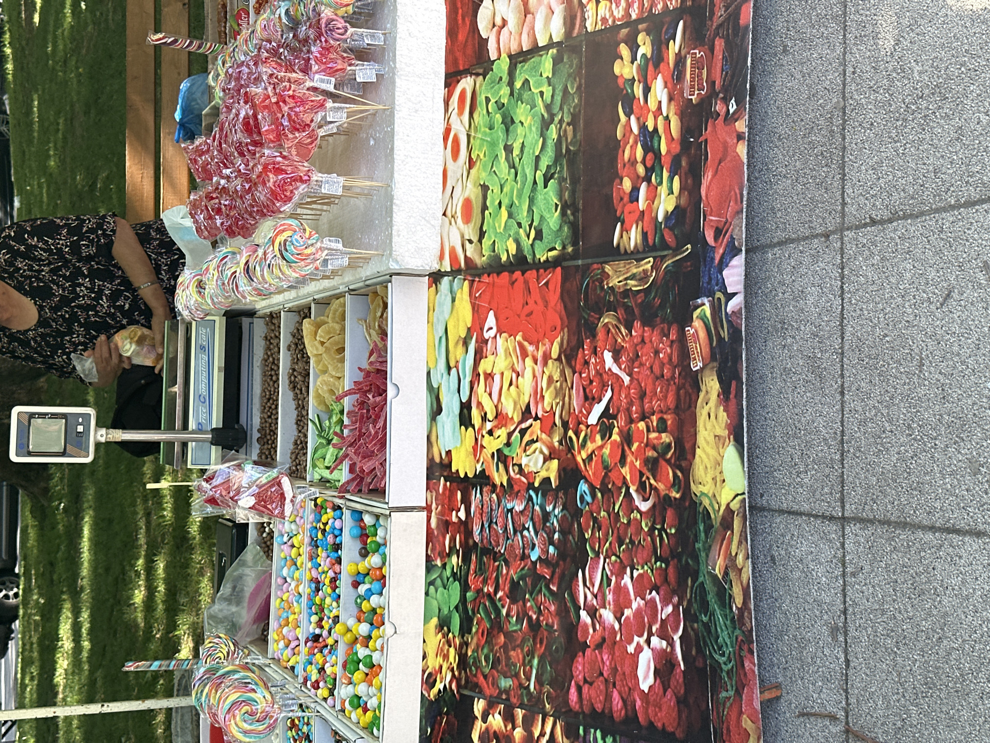 The image depicts a vendor selling candies, lollipops, and various sweets from a stall. A person is visible on the left side of the image, potentially a customer or the vendor themselves. The stall is filled with various sweets. A digital scale is on the counter. The background shows a grassy area. The location is identified as Montana, Bulgaria. The right side of the image shows the pavement.