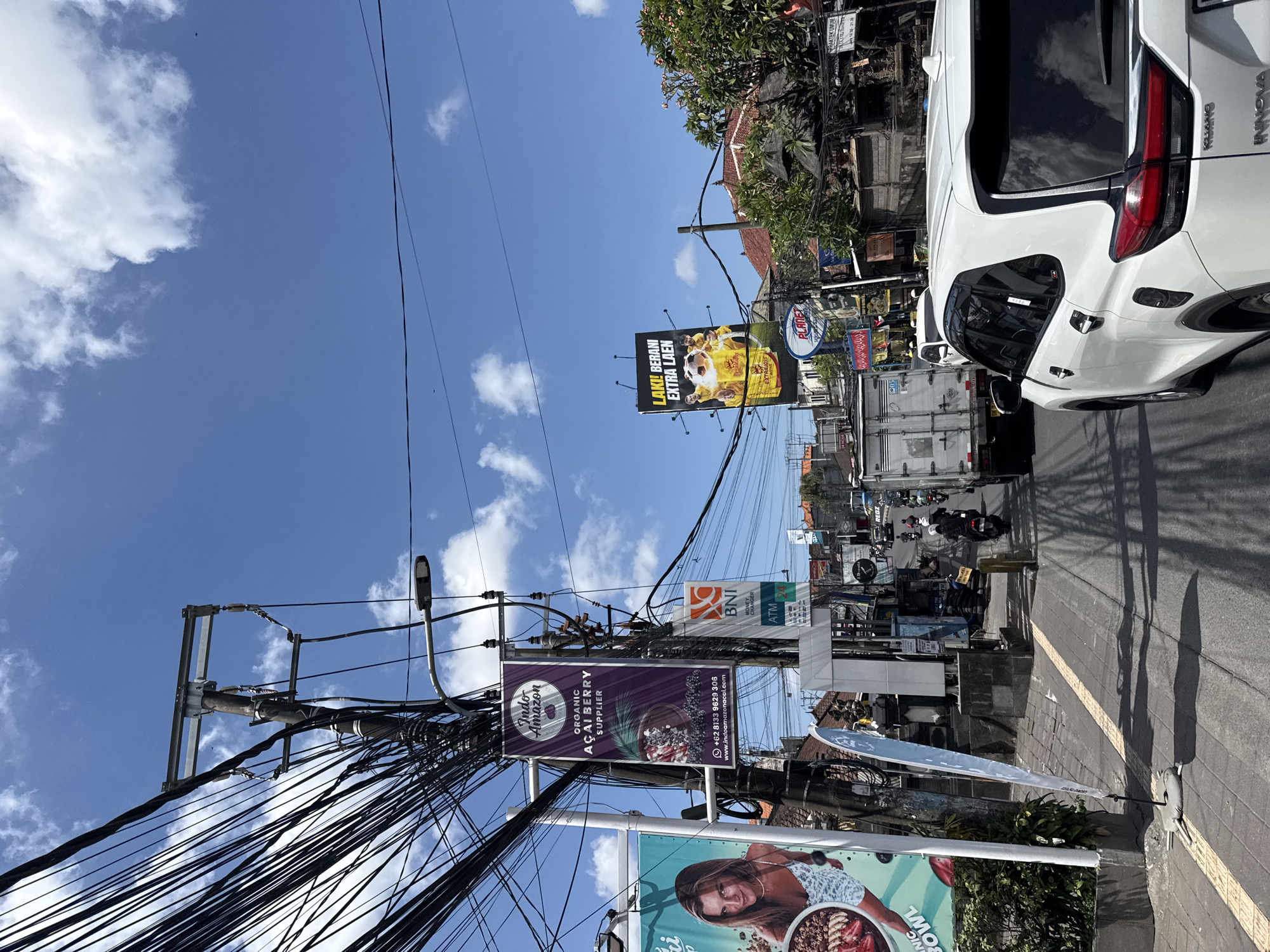 The image shows a street scene in North Kuta, Indonesia, under a clear blue sky with scattered white clouds. In the foreground, a white SUV is driving on a paved road. The street is lined with buildings and shops, many with advertising banners and signs. A prominent billboard displays an advertisement for "Indo Amazon" featuring organic acai berry supplier information and contact details. Another billboard advertises something with the text "LAKI! BERANI EXTRA LAEN".
Overhead, a dense network of electrical wires crisscrosses the sky, supported by utility poles. Traffic, including motorcycles and a white truck, is visible on the road. The overall impression is a busy, commercial street in an urban environment.
