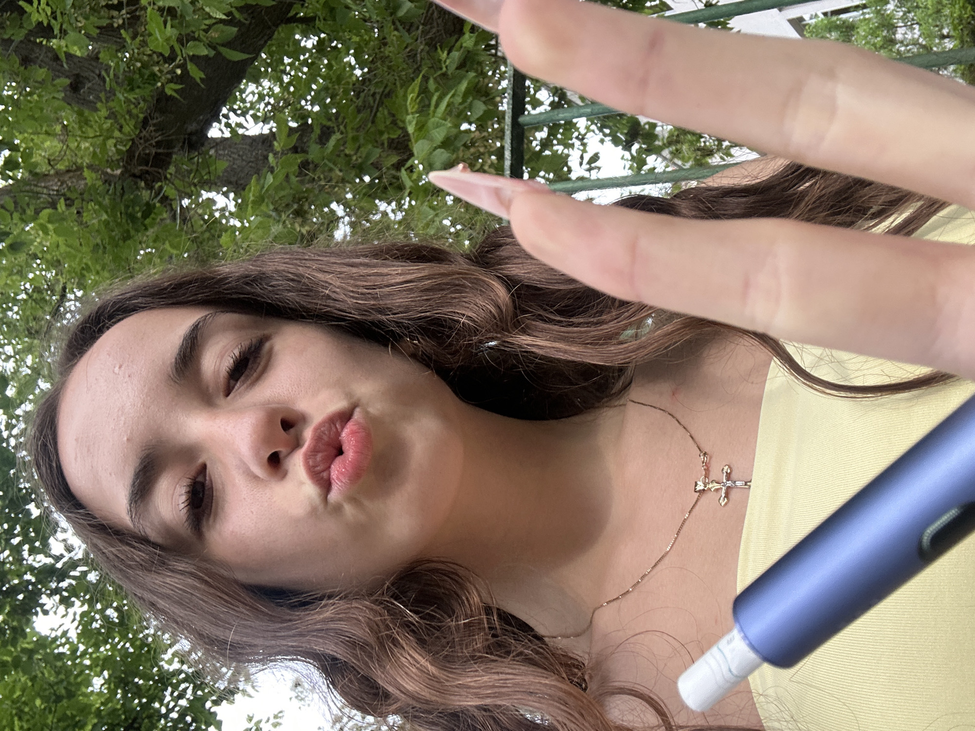 An extreme close-up selfie shows a young woman outdoors, with green foliage and trees in the background. She is wearing a light yellow top and a delicate gold necklace with a cross pendant. Her hair is long and wavy, with brown and auburn tones. Her lips are pursed in a kiss-like expression, and she is holding a dark blue electronic device in her right hand, with her fingers and long, light-colored fingernails prominently featured in the foreground. The setting suggests a park or garden, possibly in a residential area, given the presence of a green metal fence. The lighting is natural, indicating daytime. The image does not contain any specific landmarks or identifying features that would place it in Shumen, Bulgaria.