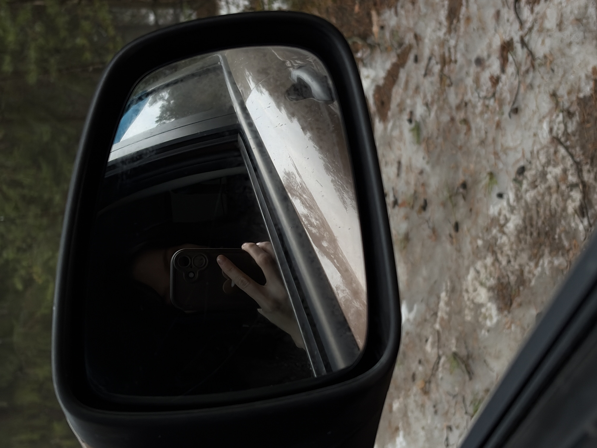 The image shows a side mirror of a vehicle reflecting the interior of the car and the ground outside.
Inside the car, a hand holding a smartphone is reflected. The phone has multiple lenses.
Outside the car, the ground is covered in a mixture of dirt, snow, and small debris like leaves and twigs. The overall environment suggests a natural, possibly wooded or unpaved area.
The location information indicates that the image was taken in Chaikovsky, Russia.