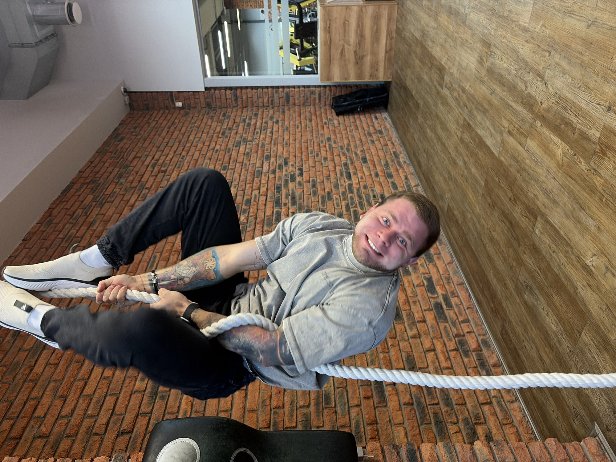 The image depicts a smiling man, likely in a gym or fitness facility in Chaikovsky, Russia, holding onto a thick white rope. He is fair-skinned with short brown hair and a beard, wearing a light grey t-shirt and dark pants. Tattoos are visible on both of his arms. He wears white athletic shoes with white socks.
The man is actively gripping the rope with both hands, suggesting he is either engaged in a climbing exercise, holding a pose, or demonstrating strength. He is looking directly at the camera with a cheerful expression.
The setting is indoors, featuring a distinctive floor made of reddish-brown brick-patterned tiles. One wall is covered in vertical wooden panels, while another is white. In the background, through a large window, yellow-framed gym equipment is visible, reinforcing the gym environment. A small, white security camera is mounted on the white wall. An overhead ventilation duct system is also visible. The lighting suggests it is daytime. No specific legible text is clearly visible on clothing or objects, though faint markings or a logo might be present on his shoe and shirt.