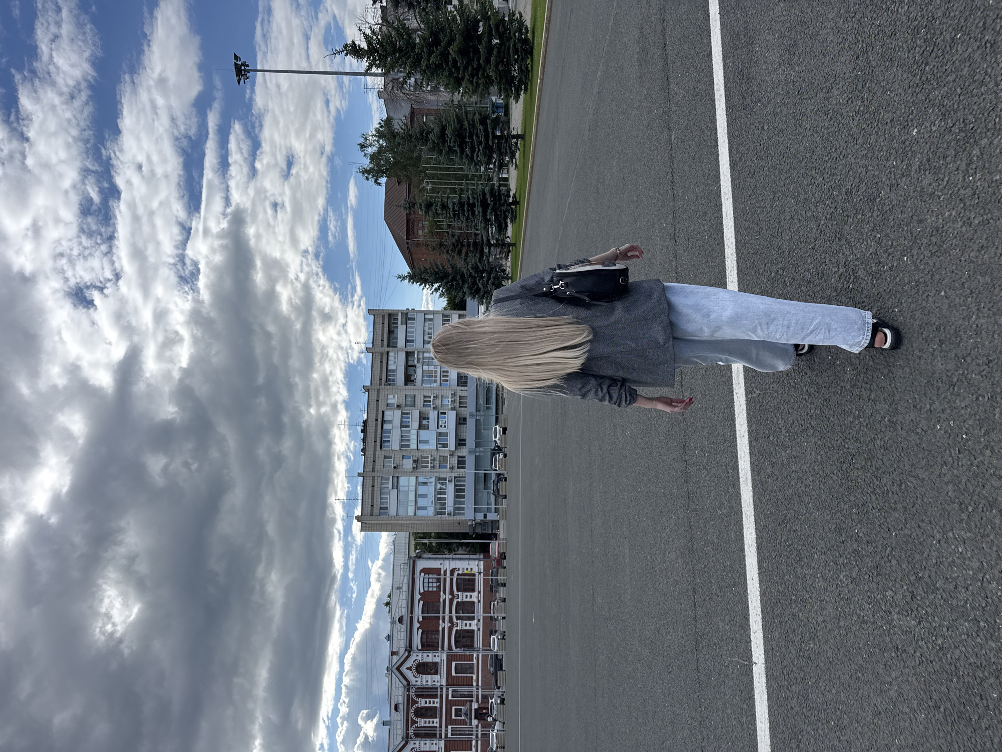 The image depicts a rear view of a blonde woman walking across a large asphalt area in the city of Novokuybyshevsk, Russia. She is wearing a grey blazer, light blue wide-leg jeans, and black sandals. A black bag is slung over her shoulder. In the background, several buildings are visible, including a red brick building with ornate white trim and a tall, multi-story residential building. Trees line the area between the asphalt and the buildings. The sky is partly cloudy, with a mix of grey and white clouds against a blue sky. A tall lamppost stands to the right of the frame. A white line is painted across the asphalt in the foreground. The scene appears to be an open public space within the city.