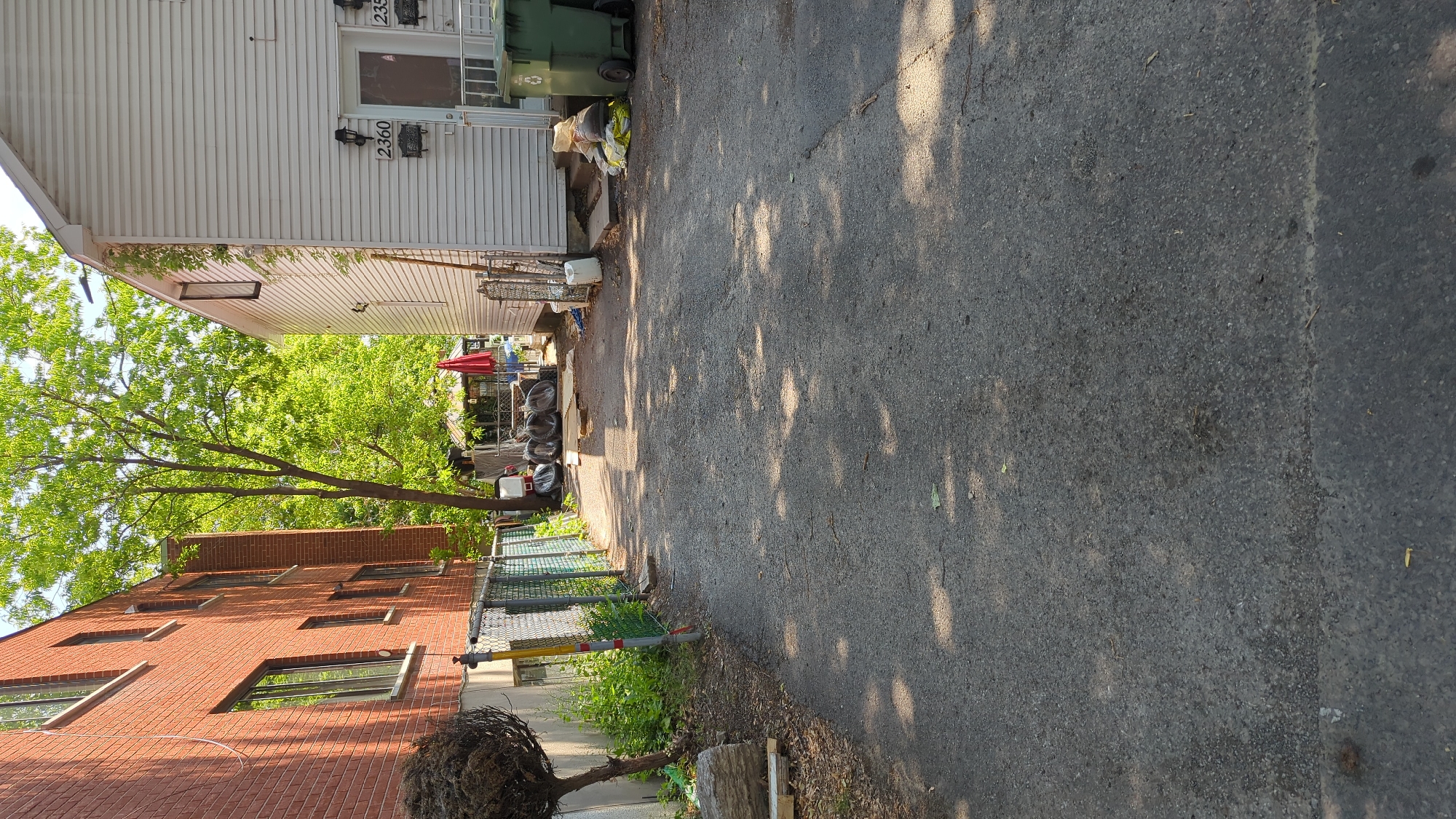 The image shows a narrow paved driveway between two residential buildings in Montreal, Canada. The building on the left is constructed of red brick and has several windows. The building on the right is clad in white siding and has two entrances visible, each with a house number: 2360 and 235... . Several large green garbage bins are situated near the entrance of the building on the right, along with piles of what appear to be construction materials or debris. In the background, there is a small gazebo-like structure with a red umbrella. The driveway is partially shaded by trees, casting dappled shadows on the pavement. There are no people visible in the image.