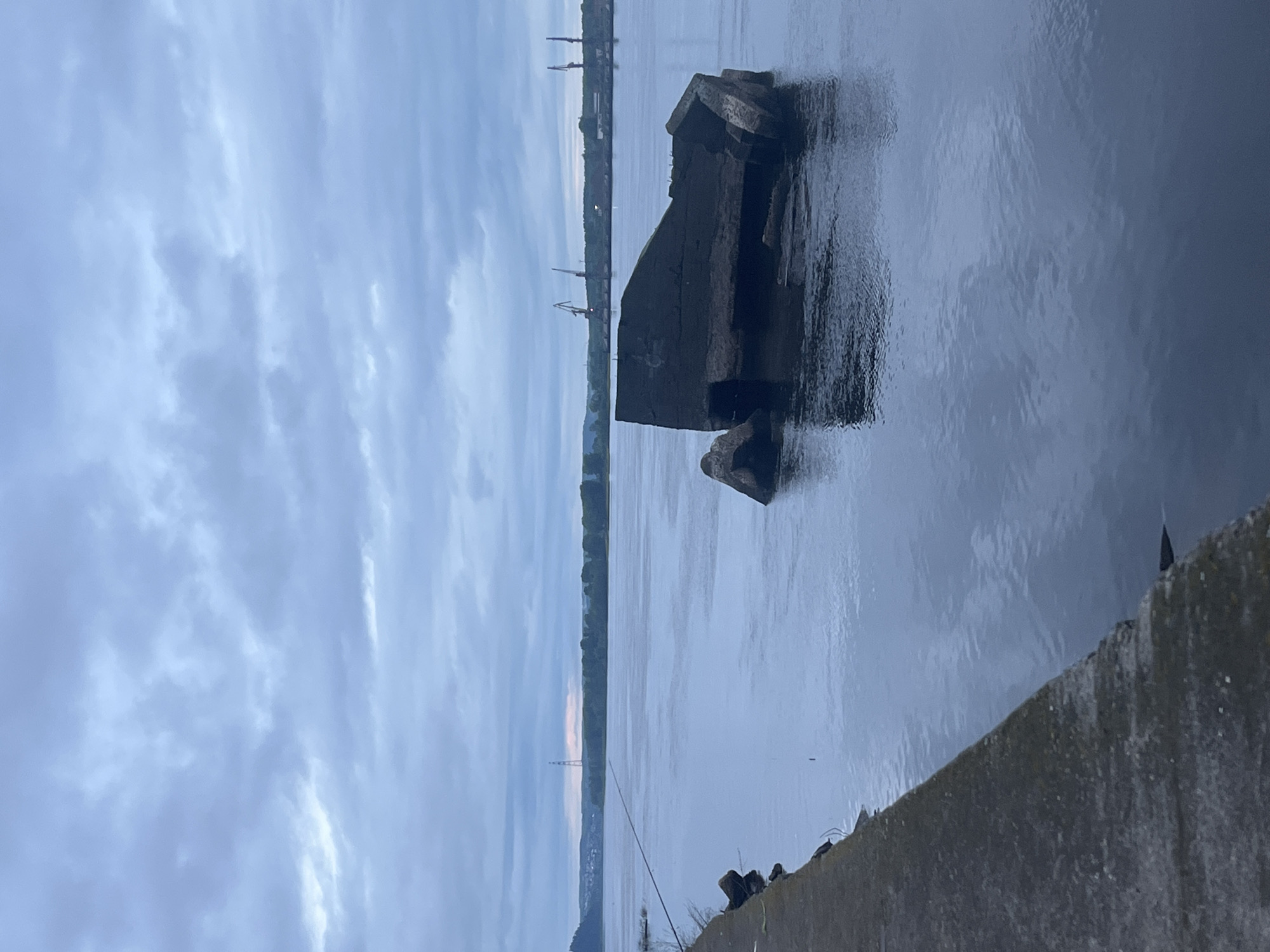 The image presents a wide, calm body of water, likely a river or reservoir, under an overcast sky in Chaikovsky, Russia. The sky is predominantly light grey-blue with scattered, diffuse clouds, suggesting either twilight or an overcast daytime condition. In the foreground, a weathered concrete embankment or pier extends diagonally from the lower left corner towards the mid-left of the frame, meeting the water's edge. A fishing rod extends over the water from the left edge of this embankment. In the midground, towards the right, a large, dark, irregularly shaped concrete structure is partially submerged in the water. Its rough, fractured surfaces are visible above the water, creating a dark reflection below. The water itself is dark blue-grey, exhibiting subtle ripples and reflecting the cloudy sky. The distant horizon reveals a low, green shoreline or forested area. On the far left of the distant shore, faint structures are visible atop a hill. Centered on the distant horizon, a tall, slender lattice tower, consistent with an electricity pylon, stands against a faint, light-pinkish glow in the sky. To the right, along the distant shoreline, several large industrial port cranes are discernible, accompanied by darker, indistinct buildings or factory structures, indicating an industrial area. No individuals are explicitly visible in the frame.