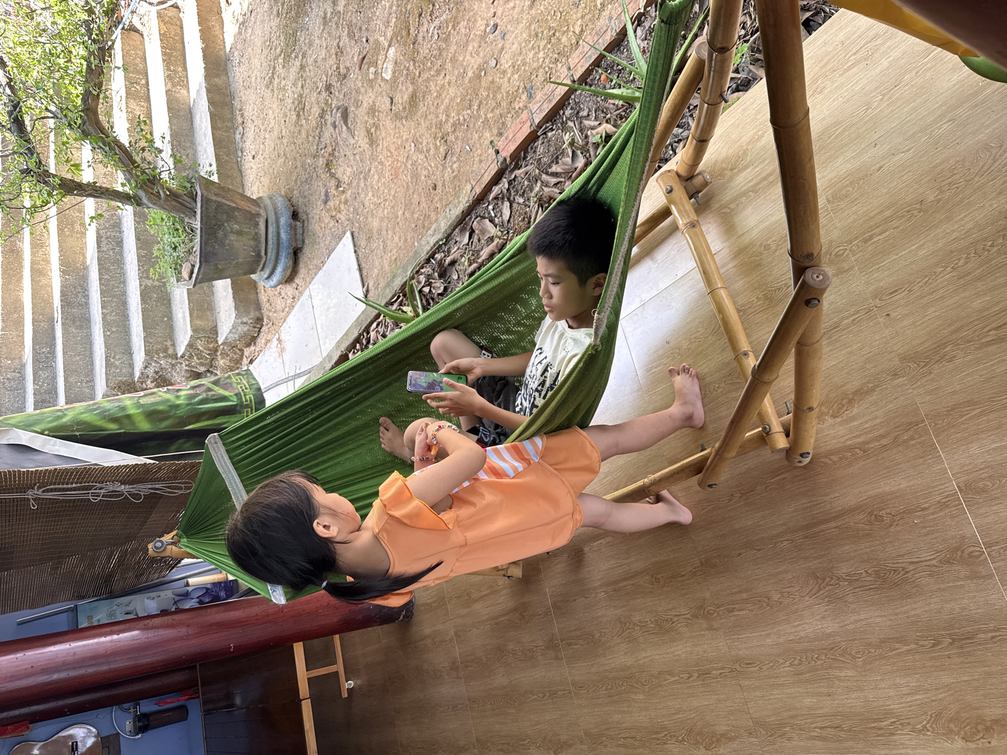The image shows two children in a green hammock inside what appears to be a home. The older child is sitting up and holding a mobile device, possibly a smartphone. The younger child is lying down, facing away from the older child. The hammock is suspended by a bamboo frame. To the left of the hammock is a dark-colored object, likely a table or shelf. In the background, steps lead to a potted plant. The floor is a light wood-grain surface. The location is identified as Tay Nha Trang Ward, Vietnam.