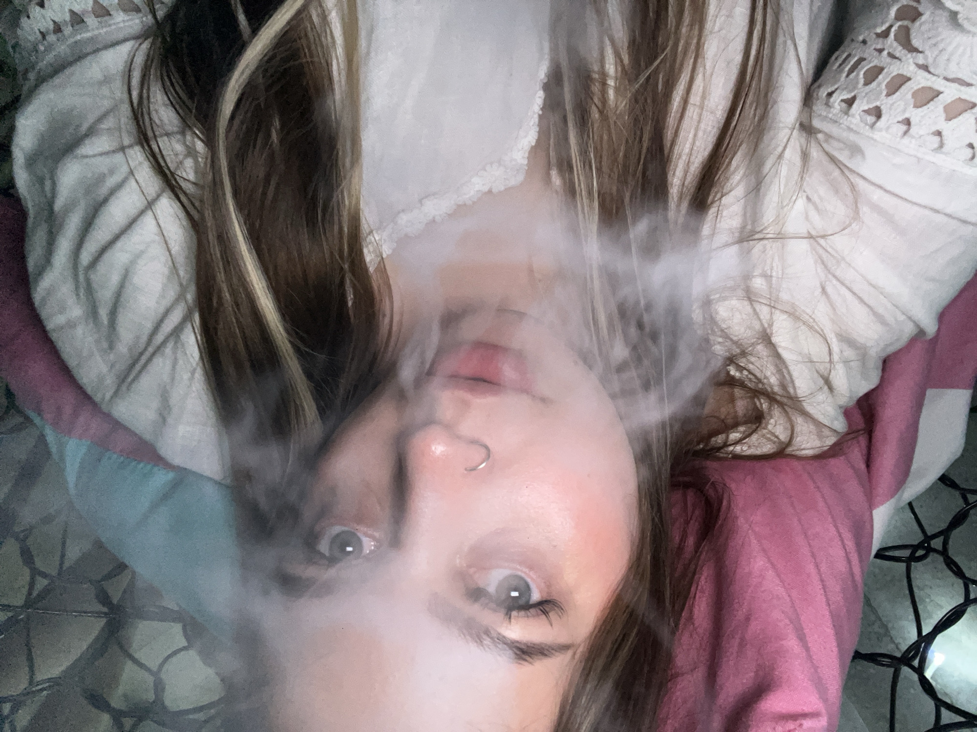 A young woman lies supine, partially obscured by a cloud of smoke. She has fair skin, light brown hair with blonde streaks, and a nose piercing. She is wearing a white, possibly cotton blouse with lace details around the neckline and sleeves. She is positioned on a surface with a pink and blue pillow, and string lights are visible in the background. The image provides no information about the city of Novopokrovskaya, Russia.