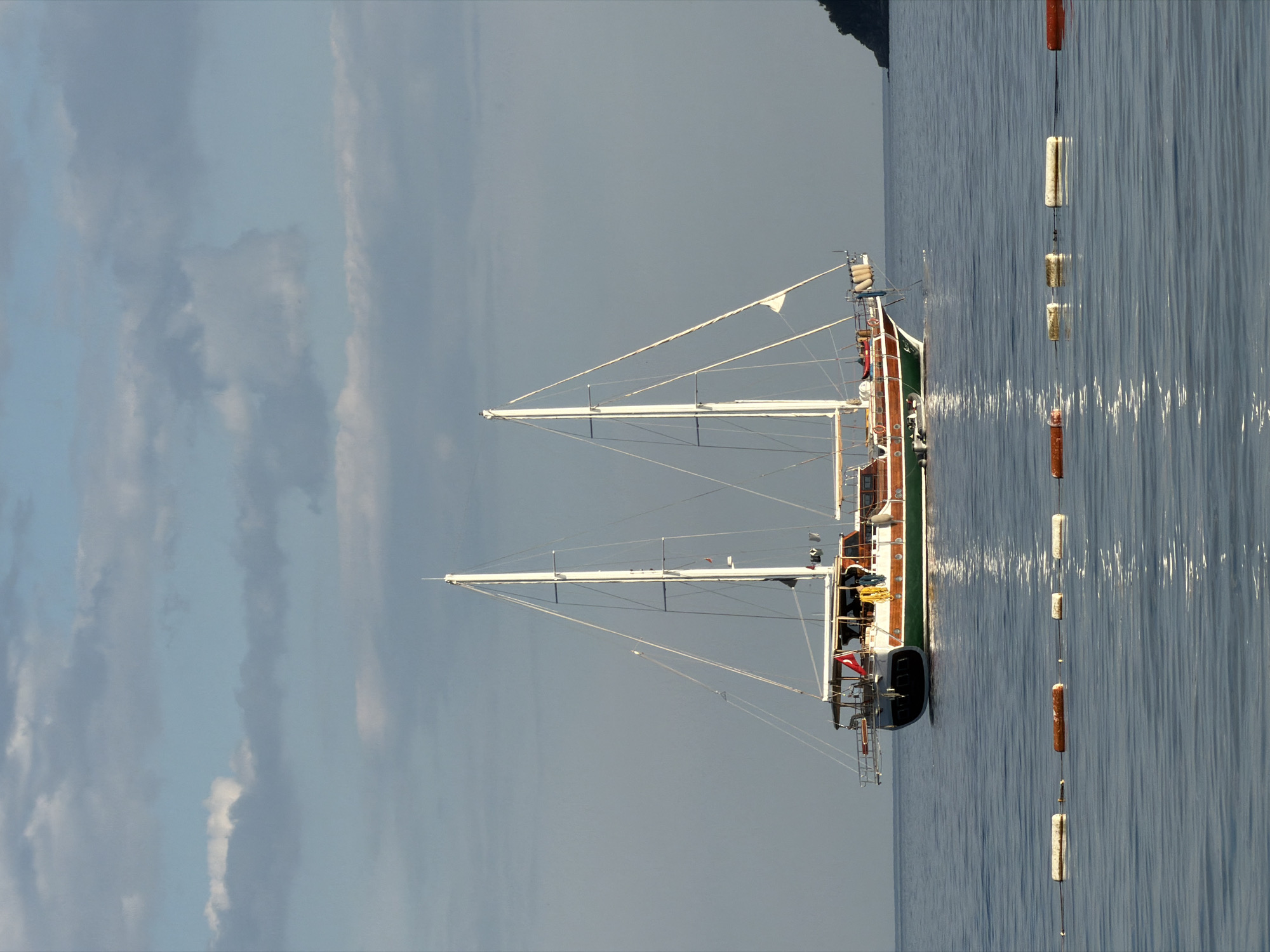 A side view of a large two-masted sailboat at rest in a body of water, likely near Budva, Montenegro, as evidenced by the presence of a Turkish flag. The boat's hull is mostly dark, with visible green and brown trim. The two masts, supporting white sails, extend high into the air. The water is a calm, reflective blue, with some ripples indicating a light breeze. Adjacent to the boat, a string of evenly spaced cylindrical buoys, alternating between white and orange colors, are suspended vertically in the water. The sky is overcast, with varying shades of gray clouds dominating the upper portion of the frame.