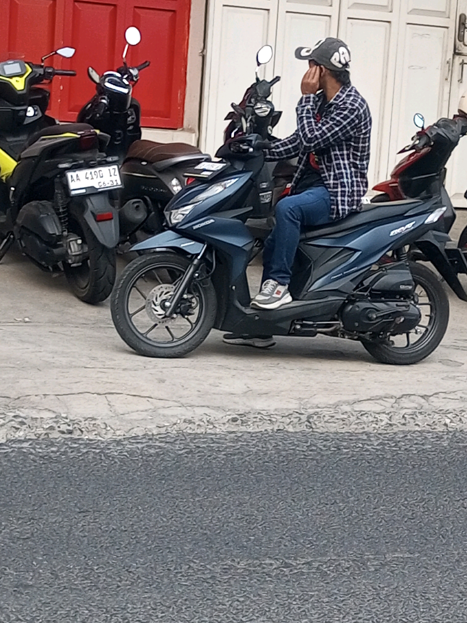 A man wearing a plaid shirt, blue jeans, a baseball cap, and sneakers is sitting on a dark blue scooter parked on a cracked concrete surface in Mojotengah, Indonesia. The man is holding his hand to his face, possibly talking on a phone. Behind him, several other parked scooters are visible, including a yellow and black scooter with a license plate reading "AA 4190 IZ". The background features a red wall and a white building with paneled doors.