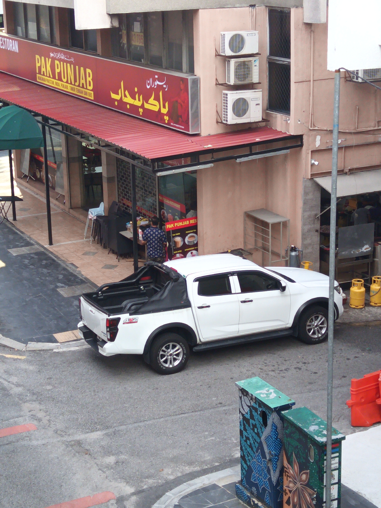 The image captures an outdoor scene in Kuala Lumpur, Malaysia, featuring a restaurant with a prominent "PAK PUNJAB" sign. The restaurant appears to be a halal establishment, as indicated by the Punjabi script and the text "PAK PUNJAB RESTAURANT" visible on a menu board. The building has a light pink facade and features several air conditioning units mounted on the wall. A white pickup truck is parked on the street in front of the restaurant. The street is paved with asphalt and bordered by concrete sidewalks. In the foreground, there are two decorative electrical boxes with artistic designs. The scene suggests a typical urban environment with commercial activity.