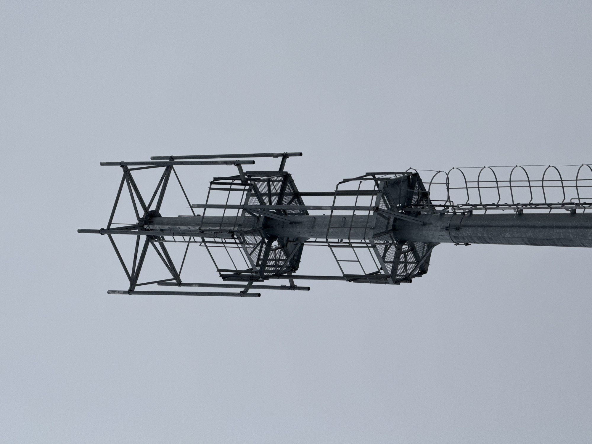 The image shows a low-angle view of a metal structure against a pale, overcast sky. The structure is likely part of a utility tower or a similar industrial installation. It consists of a central cylindrical element with latticed platforms extending from it. The platforms are constructed from metal beams and gridded panels. A cage-like safety enclosure runs along the top of the structure. The absence of identifying marks or context makes it difficult to ascertain the specific location or purpose of the structure within Kirovsk, Russia. No people or animals are visible in the image. The environment appears to be uniformly grey, suggesting a dull, cloud-covered day.