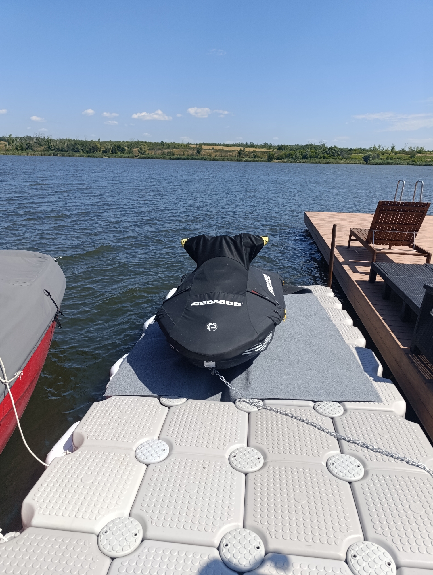 A jet ski is covered with a black tarp and parked on a modular floating dock on a body of water. The dock is made of interlocking white plastic modules. To the left, a portion of a red boat covered with a gray tarp is visible. In the background, a wooden deck with a lounge chair and a table is situated on the shore, which is lined with trees and vegetation under a clear blue sky with scattered white clouds. The location is the Dnipro River in Bulatselivka, Ukraine.