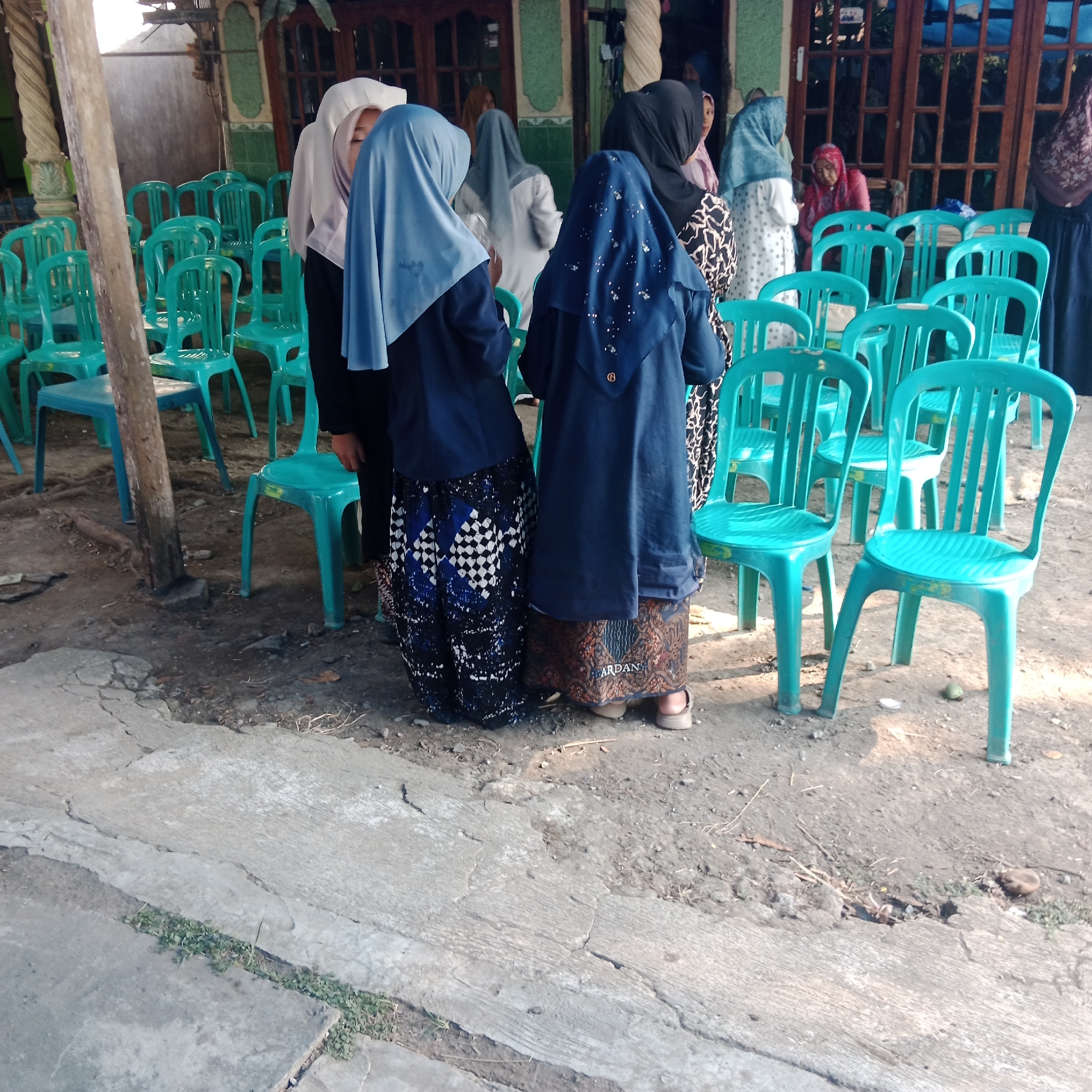 This image depicts an outdoor gathering in what appears to be the city of Tegowanu Kulon, Indonesia. In the foreground, two individuals are seen from the back, both wearing dark blue outer garments and patterned skirts. They are standing on a cracked concrete surface. Behind them, rows of bright teal plastic chairs are arranged, suggesting a seated event or waiting area. The background reveals a building with wooden-framed windows and a green tiled facade. Several other individuals, mostly women, are visible in the background, also wearing headscarves and modest clothing, some standing and interacting. The overall atmosphere suggests a community event, possibly religious or social, taking place in a semi-public outdoor space adjacent to a building. The ground is a mix of concrete and dirt with scattered debris.