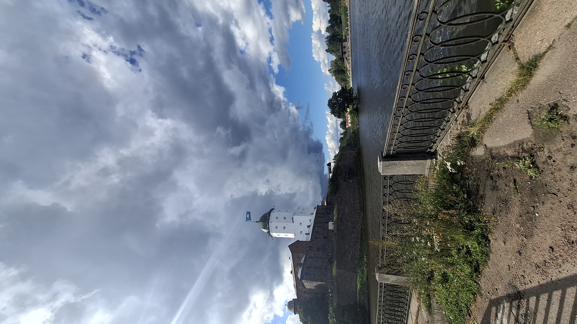 A vertical, outdoor, daytime photograph shows Vyborg Castle, a prominent landmark in Vyborg, Russia, situated on a hill overlooking a body of water. The sky is dramatic, with a mix of dark, heavy clouds and patches of bright blue sky with white cumulus clouds.
The castle complex consists of stone fortifications and a distinctive white, round tower topped with a dark green roof. A blue and yellow flag flies from the top of the tower. The castle is built on a stone embankment that separates it from the water.
In the foreground, a dark metal railing with an ornate, oval design runs along a concrete pathway. This pathway is partially covered with soil and patches of wild greenery, including tall grasses and small white flowers. The water is dark and appears choppy, reflecting the moody sky. Across the water, the opposite bank is lined with trees and some distant buildings, suggesting a cityscape beyond the natural elements. The overall scene conveys a sense of historical grandeur set against a dynamic natural backdrop.
