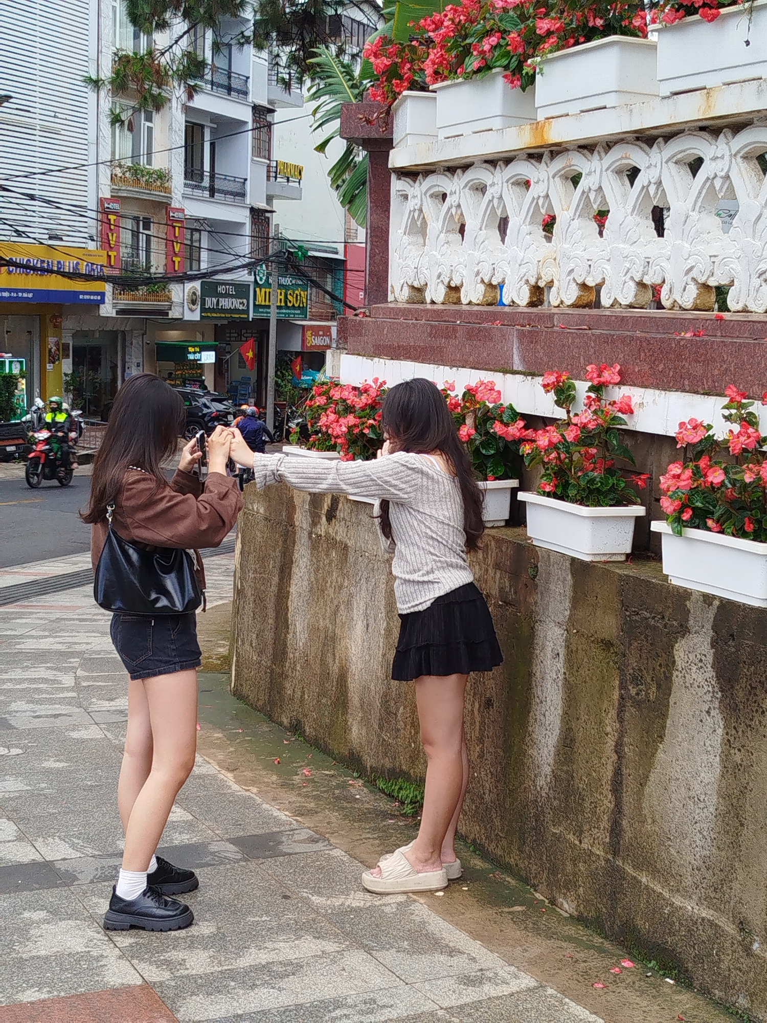 Two young women are interacting on a sidewalk in Da Lat, Vietnam. One woman, wearing a brown jacket, dark denim shorts, white socks, and black shoes, holds a smartphone and appears to be photographing the other woman. The second woman wears a light grey long-sleeved top, a short black skirt, and light-colored sandals. She stands with her back to a concrete wall adorned with white planters filled with vibrant red flowers. The wall has a decorative white railing above the planters. In the background, street scenes include parked cars, motorbikes, and commercial establishments with signs in Vietnamese such as "DUY PHUONG 2" and "MI H SON," indicating hotels and restaurants. The architecture features French colonial-style buildings common in Da Lat. The day appears overcast.