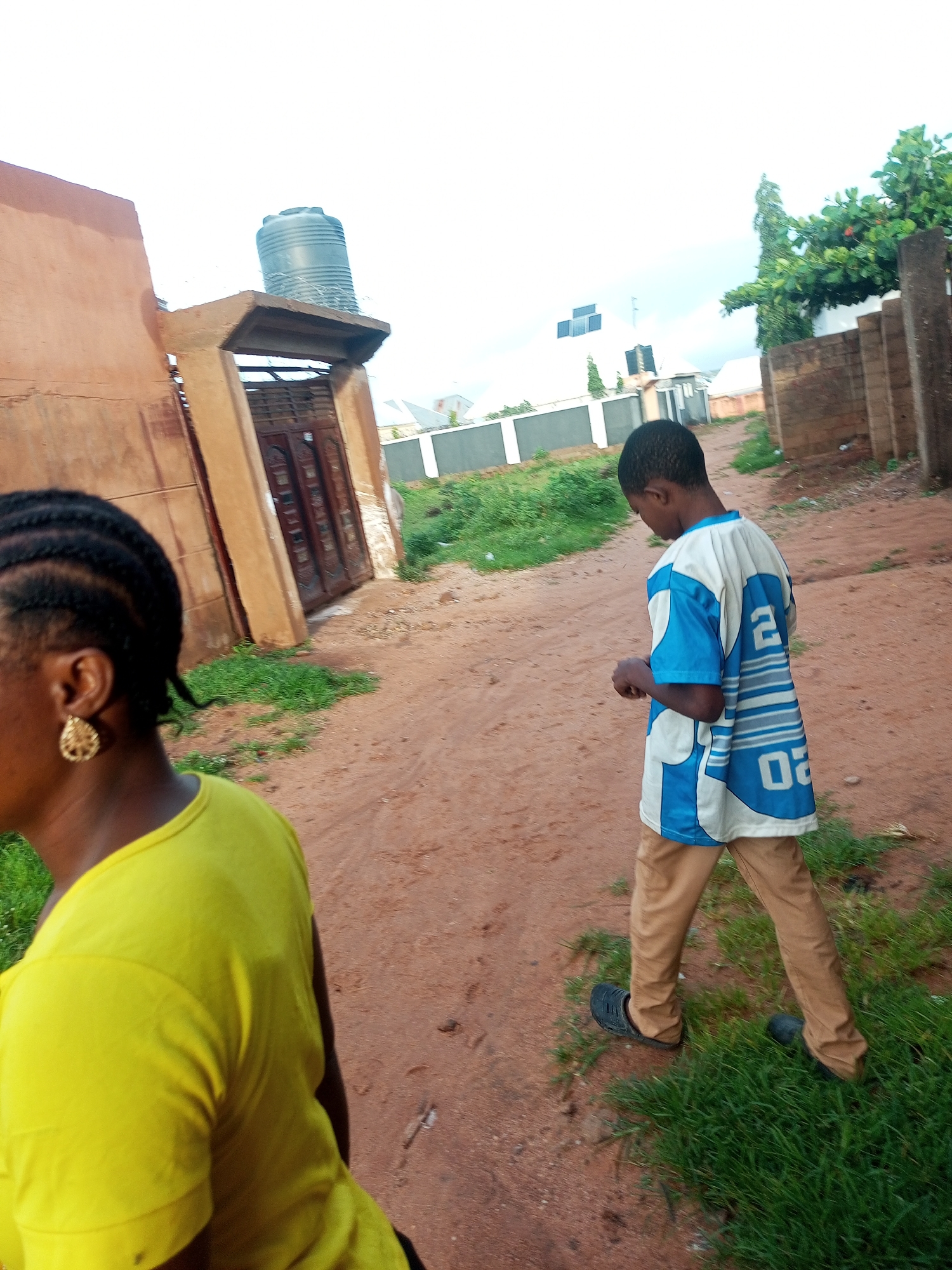 In Tempure, Nigeria, a young boy wearing a blue and white shirt and tan pants walks on a dirt path next to a woman with braided hair and a yellow shirt. The scene appears to be a residential area with buildings and vegetation. A large black water tank sits atop a structure to the left, and a fence encloses a white building in the background. The sky is overcast, and the overall lighting suggests it is daytime.