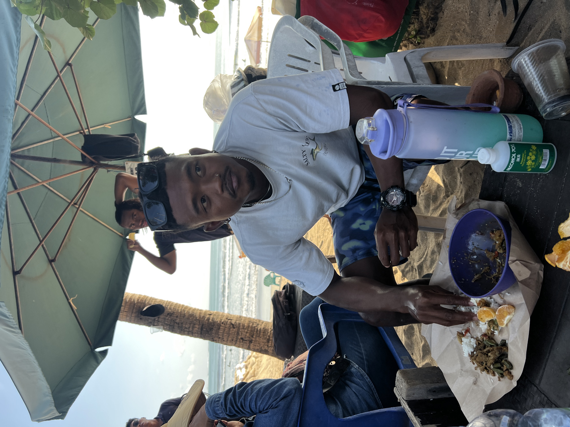 In Kuta, Indonesia, a man sits at a table on a sandy beach under a large green umbrella. He wears a white t-shirt with a shark graphic and patterned shorts, and has sunglasses perched on his head. In front of him, on brown paper, is a meal with rice, vegetables, and slices of orange. He is using his hands to eat. A purple bowl sits to the side, also containing food. Nearby are a large water bottle, a small bottle labeled "ALCOHOL 70% IKA", and a clear plastic cup. In the background, the ocean with gentle waves is visible, along with a palm tree and other beachgoers. Two other men are partially visible, one holding the umbrella and another in the background looking away.