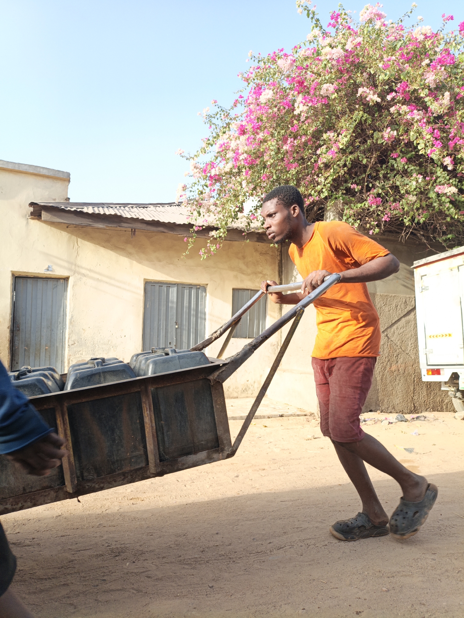 In Gwagwarwa, Nigeria, a young man in an orange shirt and reddish shorts is pulling a heavily loaded, dark-colored metal cart down a dirt road. The cart contains several black, rectangular containers with lids. The man is wearing dark-colored sandals and appears to be straining with the weight.
The background includes a single-story, light-colored building with two metal doors and window, and a white truck with a logo partially visible. A large bougainvillea bush with pink and white flowers is also present in the background. The sky is a clear, light blue.
