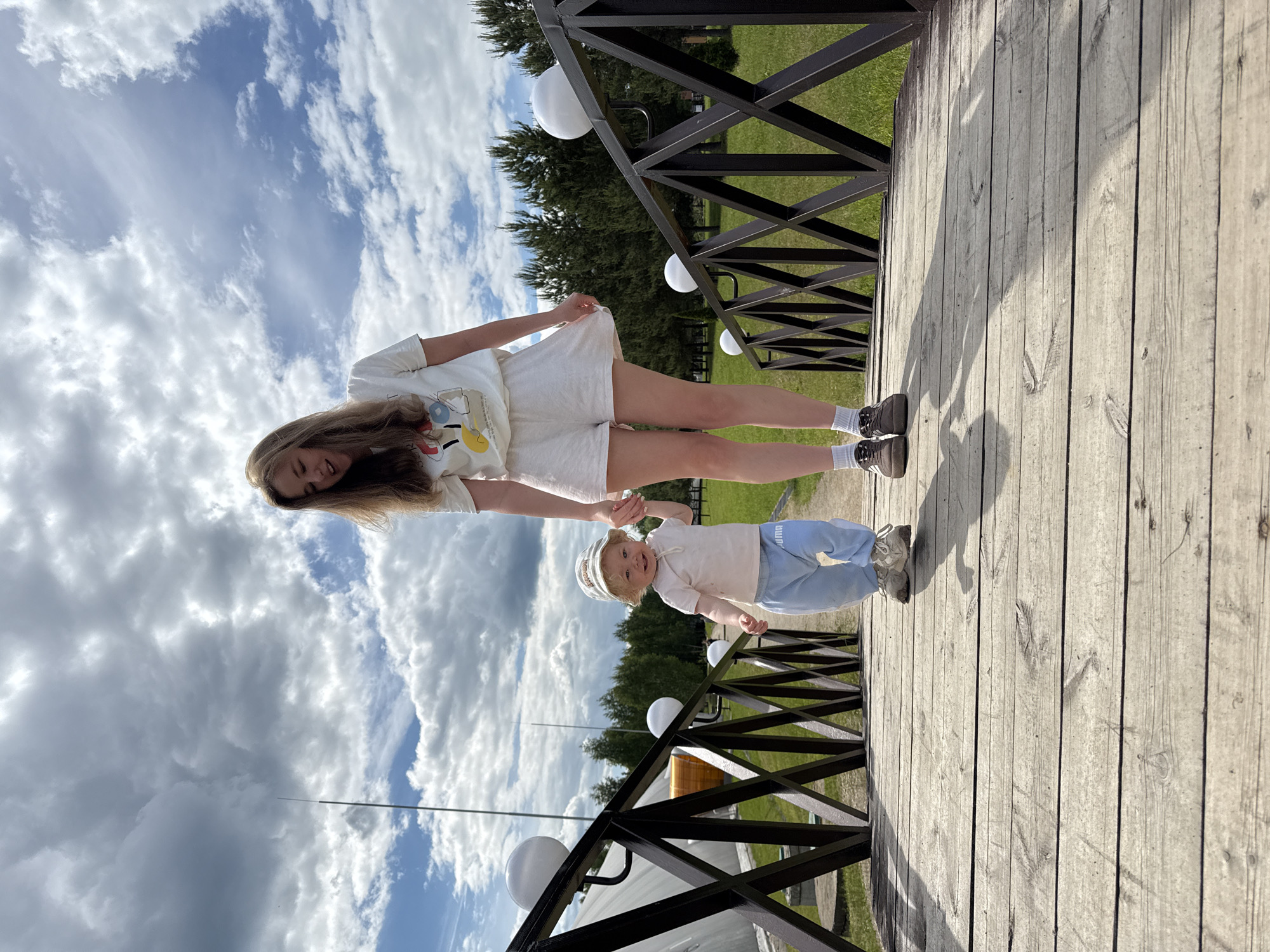 The image portrays a heartwarming outdoor scene featuring a young woman and a toddler on a wooden walkway in Dobrograd, Russia. The woman, with long, light brown hair, is dressed in a white t-shirt with a colorful graphic design, white shorts, white socks, and dark sneakers. She smiles gently while holding the hand of the young child.
The toddler, with light-colored hair and wearing a light-colored cap, a white t-shirt, and light blue pants with the text "PUMA" visible on one leg, is also smiling broadly and looking towards the camera. The child wears light-colored sneakers.
They are standing on a wooden plank walkway that appears to be part of a bridge or elevated path. A dark-colored, possibly metal, railing runs along the side of the walkway, featuring several white spherical decorative lights. In the background, there is lush green grass and dense coniferous trees under a bright blue sky dotted with scattered white clouds. The time of day appears to be sunny daylight, casting distinct shadows on the wooden surface. The shared smiles and hand-holding between the woman and child convey a joyful and affectionate interaction.