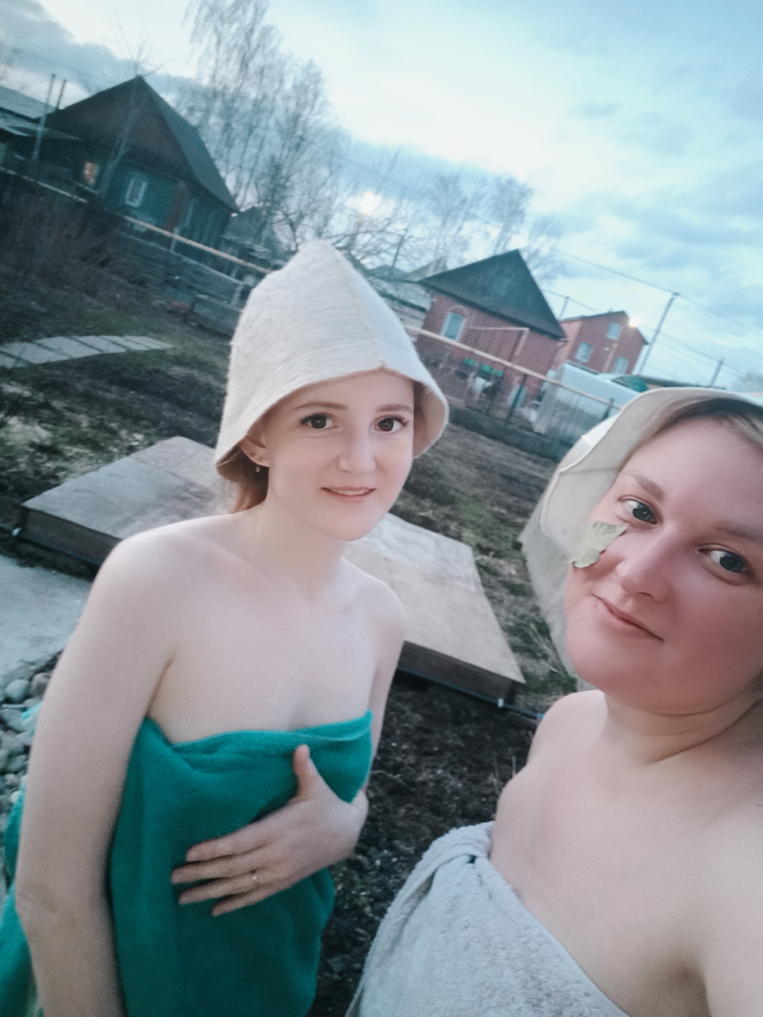 Two women are captured in an outdoor setting, likely in Chaikovsky, Russia, after or during a traditional banya (sauna) session. Both are wearing light-colored, conical felt sauna hats and are wrapped in towels.
The woman on the left, with reddish-blonde hair, smiles directly at the camera. She is wrapped in a teal-green towel, securing it with her right hand, where a ring is visible on her ring finger. Her left shoulder is bare. The woman on the right, with blonde hair, is partially visible and has a small green leaf resting on her right cheek. She wears a lighter, possibly white or light grey, towel and looks towards the camera with a gentle, content expression.
The background features a rural or suburban landscape with several wooden houses, some painted in darker colors, under an overcast sky. Bare trees suggest late autumn, winter, or early spring. The ground is a mix of grass and dirt, with stepping stones forming a pathway and wooden plank structures visible. The light suggests it is daytime, possibly late afternoon or early evening, under cloudy conditions. No visible text is present in the image.