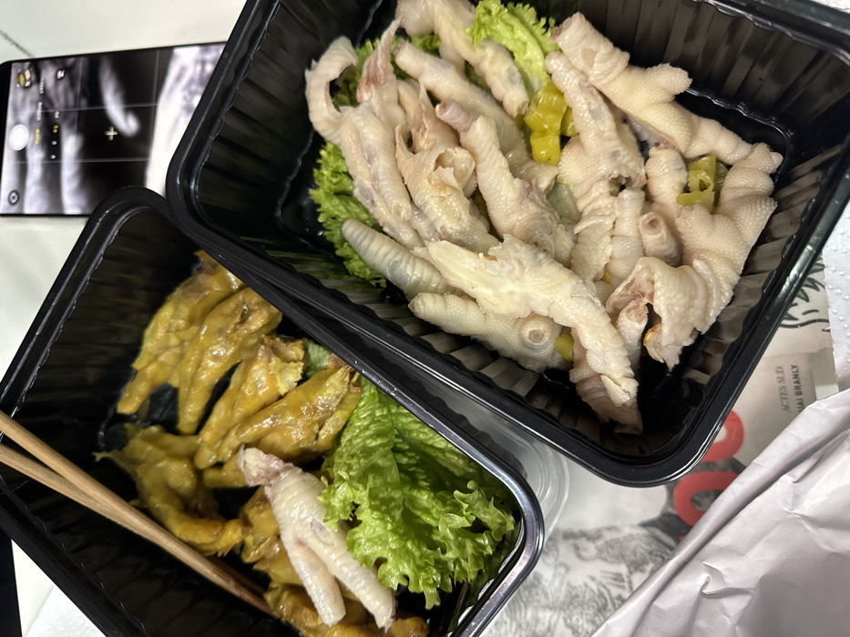The image displays two black plastic food containers filled with chicken feet, placed on a light-colored surface, possibly a table. To the left, a smartphone with its camera interface visible is lying face up on the same surface.
The container on the top right holds a generous portion of light-colored, likely blanched or boiled, chicken feet. These chicken feet are pale white to light yellow, and some small pieces of green chili or bell pepper are interspersed among them. A bed of fresh green lettuce is visible underneath the chicken feet, providing a color contrast.
The container on the bottom left contains chicken feet that are distinctly yellowish-brown, suggesting they have been marinated, braised, or stewed in a sauce. Similar to the other container, there is some green lettuce visible beneath these chicken feet. A single, lighter-colored chicken foot also appears in this container, near a pair of wooden chopsticks that rest partly inside.
The smartphone in the upper left corner shows a screen with a camera application open. Elements like "PHOTO" and "VIDEO" modes, zoom options ("1x", "0.5x"), and focus squares are discernible, indicating it is actively in photo-taking mode. The phone's screen reflects parts of the surrounding environment, including a hand or arm.
To the far right, a crumpled white paper bag or wrapper is partially visible. On this paper, some faint text can be seen, which appears to include "ACIES SUD" and "HAI BRANLY", possibly from a product label or packaging. The overall setting appears casual, likely indoors, and suggests a meal, possibly takeout or delivery, about to be consumed.