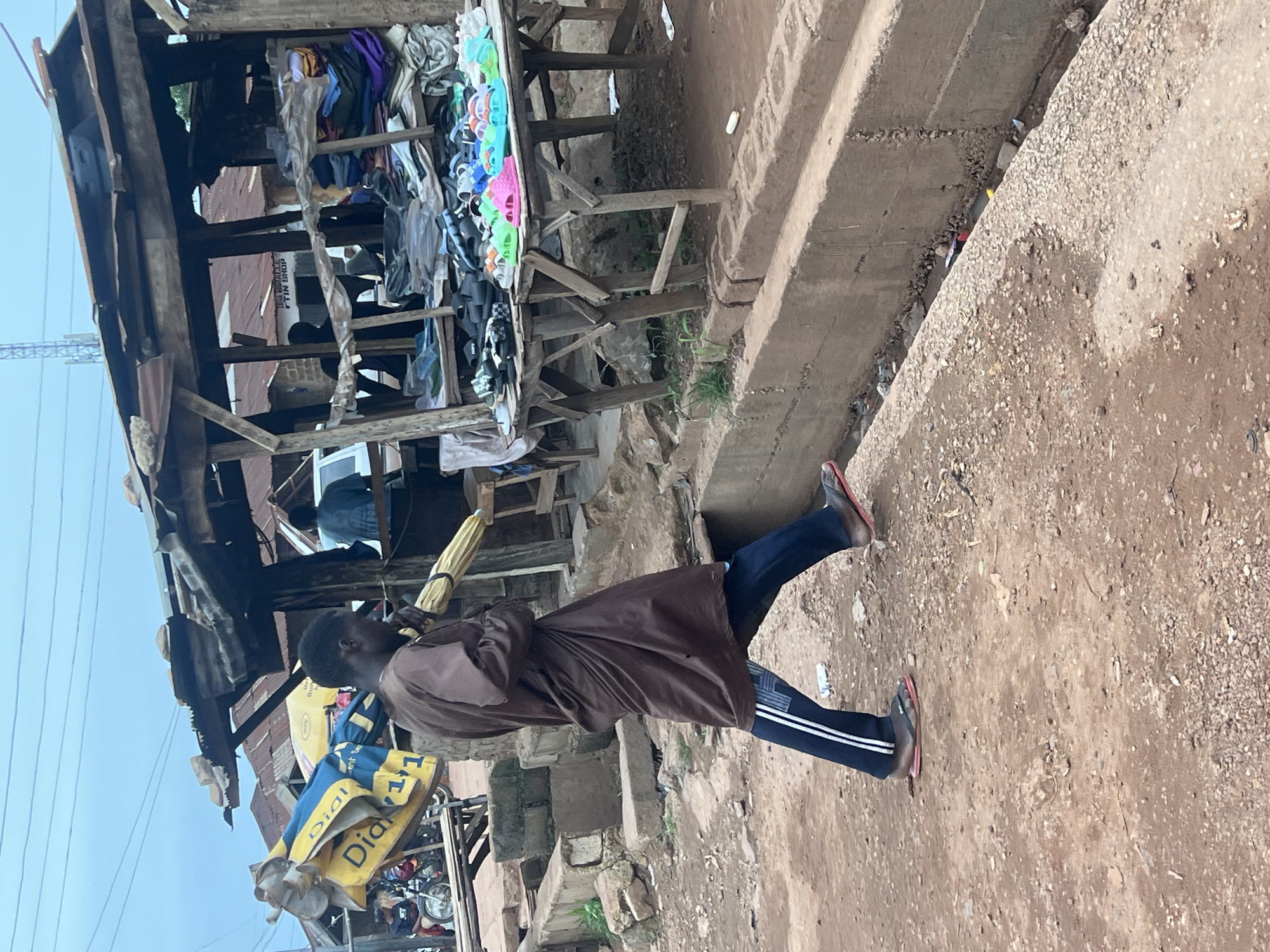 In Durumi, Nigeria, a man dressed in a brown, flowing garment and dark trousers with white stripes walks along a dirt path, carrying a large, yellow umbrella or rolled-up mat over his right shoulder. His left leg is extended forward, and he wears red-soled flip-flops. He is walking past a makeshift market stall constructed with wood and corrugated metal roofing, which is partially collapsed and weathered. The stall displays various items, including folded clothes and rows of colorful sandals and flip-flops on wooden tables. Behind the stall, other similar structures and a white vehicle are visible. The ground is predominantly reddish-brown dirt with scattered rocks and debris. A concrete drainage ditch runs alongside the path, containing some trash. The sky is overcast.