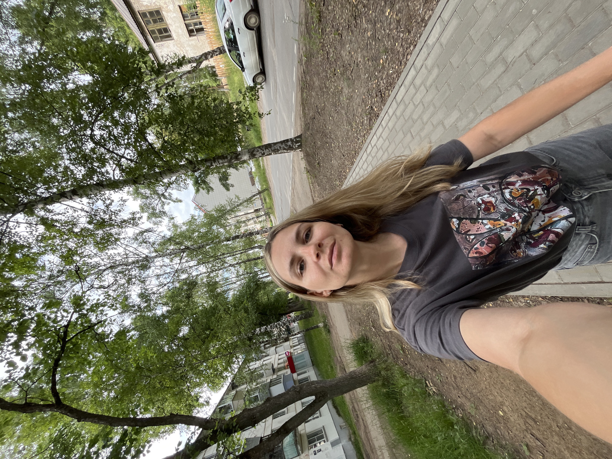 The media file is a selfie taken outdoors in Chaikovsky, Russia. The subject is a young woman with long blonde hair, wearing a dark gray t-shirt with a graphic print and dark jeans. She is looking directly at the camera with a slight smile.
The setting appears to be a residential street with trees lining a sidewalk. In the background, there are apartment buildings and a street with a white car parked on the side. The time of day seems to be daytime, with overcast skies suggesting possibly cloudy weather. The overall atmosphere is casual and everyday. There is no visible text in the image.