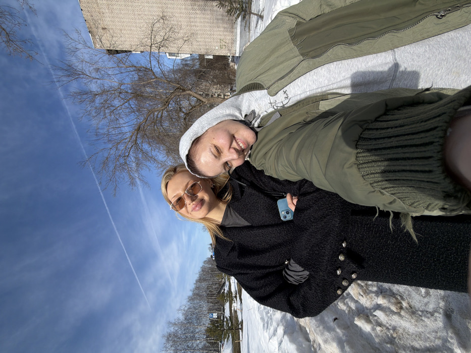 This image captures a selfie of two young adult females taken outdoors on a sunny day in Chaikovsky, Russia. The scene is set in late winter or early spring, characterized by patches of melting snow on the ground and bare deciduous trees. A clear blue sky, marked by a single white contrail, is visible overhead.
The subject on the left has blonde hair, wears light brown-tinted sunglasses, and a textured black long coat adorned with multiple silver-colored buttons on the cuffs. She holds a light blue smartphone in her left hand, tucked into the coat's opening. Her expression is a faint smile. The subject on the right, who appears to be holding the camera, wears a grey hooded sweatshirt under an olive green bomber jacket. A partial inscription, "Fight 2x," is visible on her sweatshirt. She smiles broadly and leans her head against the left subject.
In the mid-ground, behind the individuals, a multi-story building constructed of light-colored bricks is present to the right, displaying the number "57" on its facade. To the left and center background, bare trees, including several birches and a small evergreen, are visible, along with a partially obscured white road sign featuring a blue "P" symbol. The overall environment suggests an urban or suburban setting.