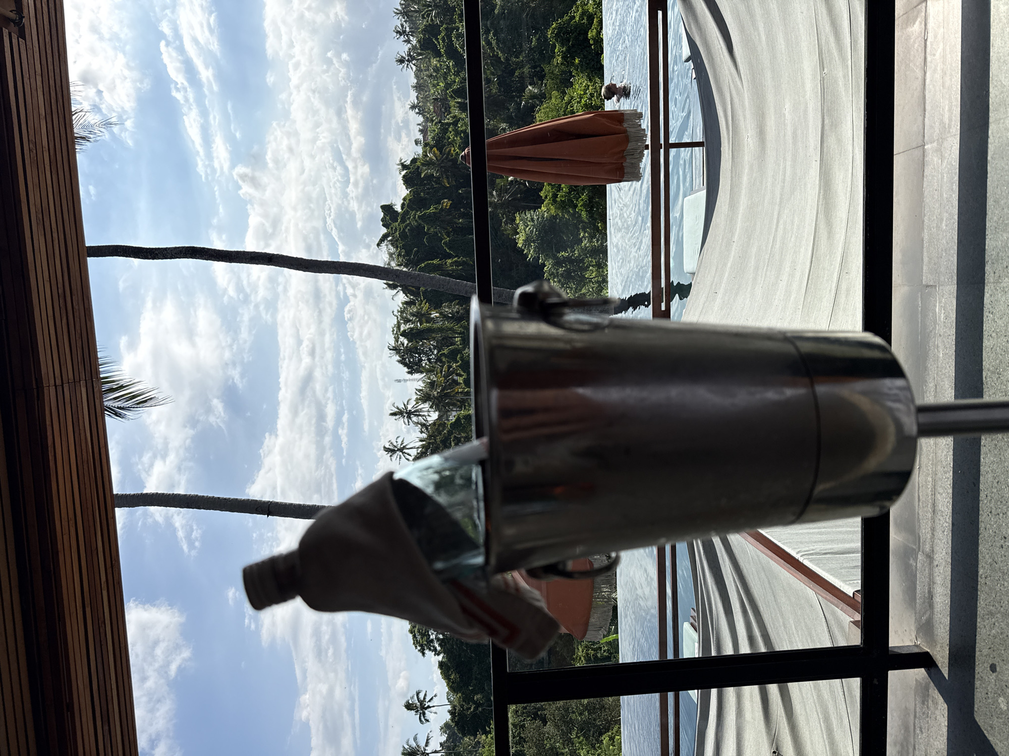 The image, taken in the Ubud District, Indonesia, shows a view from a likely elevated location with a lush green tropical forest visible in the background. Part of a swimming pool and a sun umbrella are visible through a glass window. In the foreground is a metal ice bucket with a bottle of wine or other beverage inside, draped with a cloth. Palm trees are visible in the background, scattered amidst the dense vegetation. A person can be partially seen in the pool. The photo has been rotated slightly, with the landscape appearing at an angle.