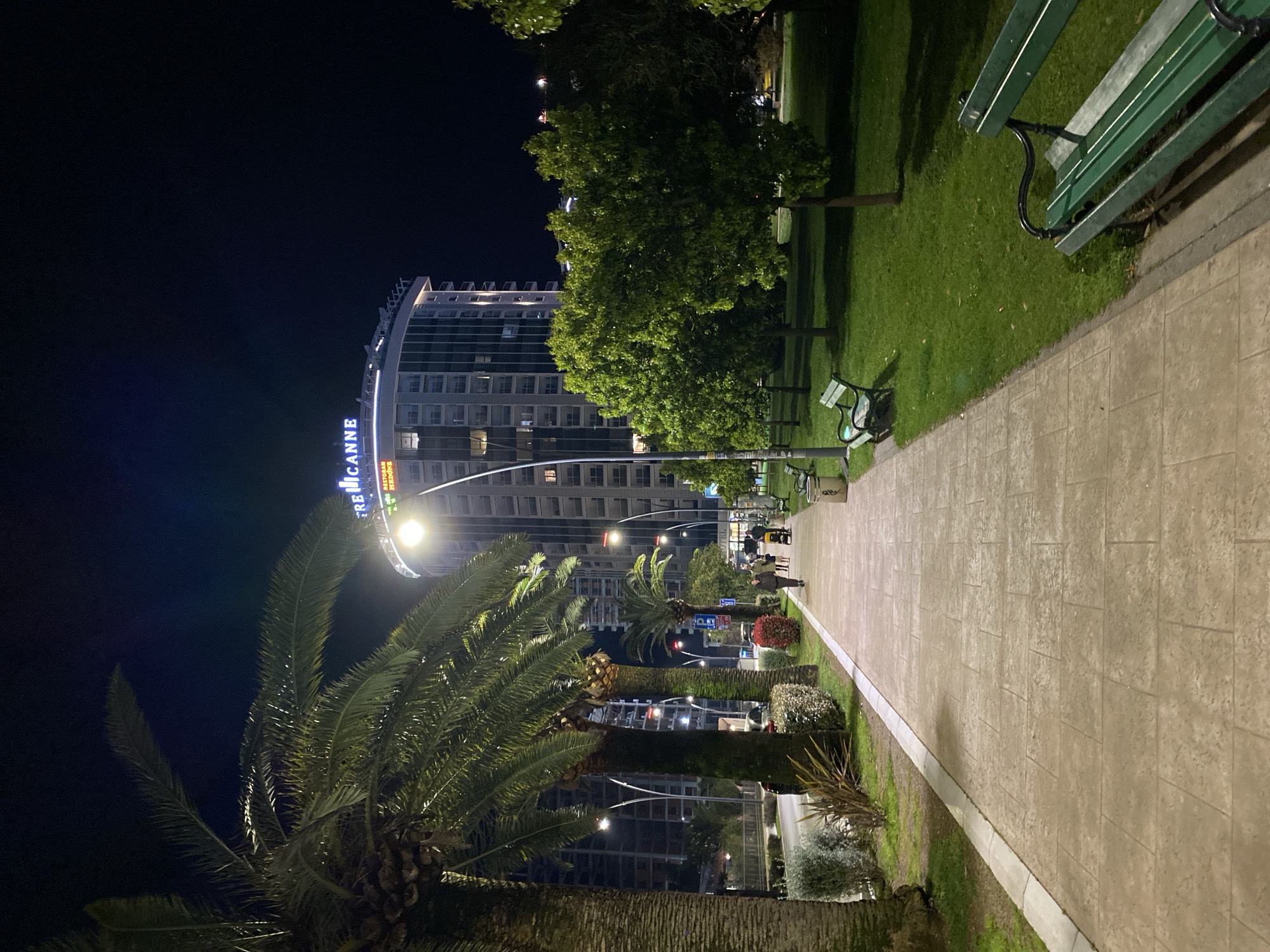 The image displays a nighttime urban scene in Budva, Montenegro, with the prominent Tre Canne building in the background. A wide, light-colored paved pedestrian walkway, constructed of rectangular tiles, extends from the lower center into the distance, illuminated by multiple tall streetlights featuring curved lamp posts. To the left of the walkway, a row of mature palm trees separates it from a parallel road that shows parked vehicles and additional ambient lighting. Several individuals are visible on the walkway; a group of three is in the midground, one of whom is pushing a baby stroller. To the right of the walkway, a well-maintained grassy park area contains several dark green park benches with black metal frames and numerous green, leafy trees. The multi-story Tre Canne building, a modern, curved structure with "TRE CANNE" illuminated in blue and "RESTORAN HEDON" in white on its rooftop signage, stands prominently against the dark sky. Other multi-story buildings, some with visible windows and balconies, are discernible behind the palm trees on the left side of the scene. The overall lighting is artificial, emanating from streetlights and building illumination, casting a soft glow on the immediate surroundings while the sky remains dark.