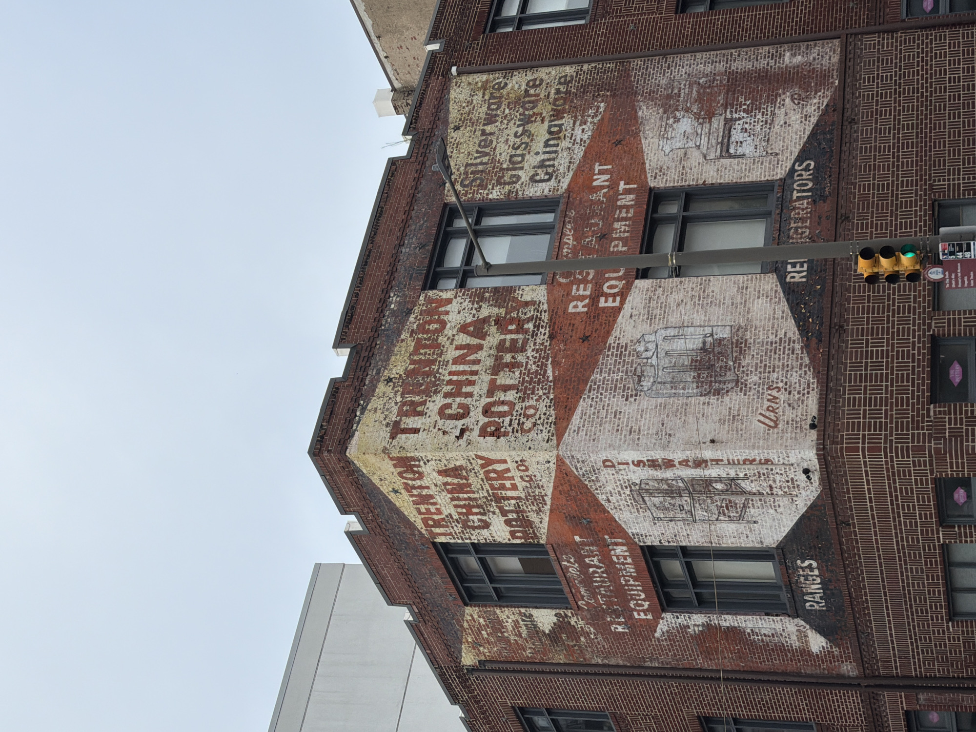 The image shows the exterior of a brick building in Philadelphia, Pennsylvania, United States of America, featuring faded, hand-painted signage. The building has multiple windows and appears to be an older structure. The signs advertise "TRENTON CHINA POTTERY CO.", "Complete RESTAURANT EQUIPMENT", "Silverware Glassware Chinaware", "RANGES", "DISHWASHERS", "URNS", and "REFRIGERATORS". The lettering is in red and black paint on a brick surface that has been painted white in some areas, with significant weathering and peeling. A traffic light with a green light illuminated is visible in the lower right quadrant of the image. The sky is overcast.