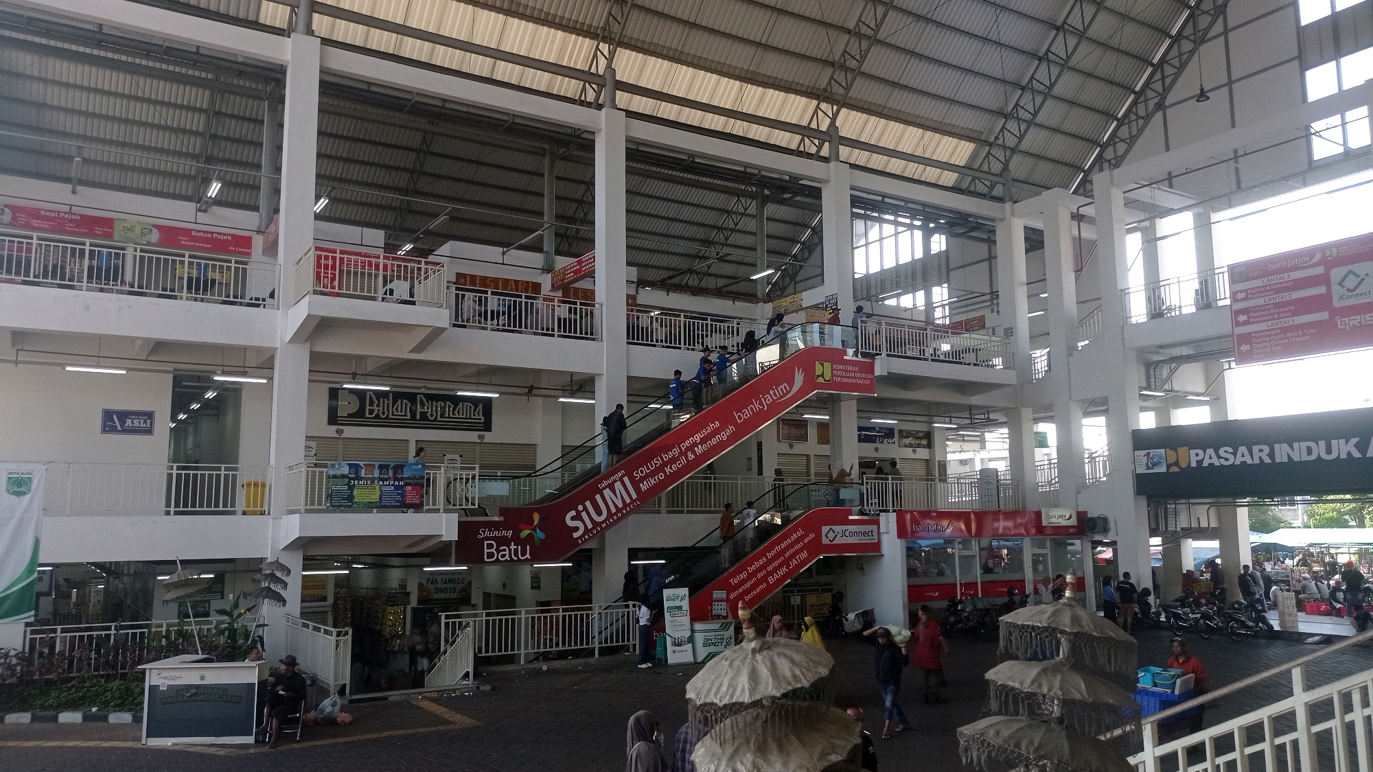 The image depicts the interior of a multi-level market building, likely a "Pasar Induk" or main market, in Batu, Indonesia. The architecture features a large, open central space with multiple levels of walkways, balconies, and staircases supported by concrete columns and a metal roof structure. Escalators, some adorned with advertisements for "bankjatim" and "SiUMI Solusi Mikro Kecil", are visible, indicating the market is a modern facility. Various shops and stalls are set up on different levels, with signage in Indonesian language. Some shops appear to be selling food, clothing, and other consumer goods.
The ground level shows a bustling environment with people walking, shopping, and interacting. Several motorcycles are parked in an open area outside the market, suggesting it is a public transport hub. People are dressed in casual attire, typical for a market setting. Some individuals are seen carrying bags, and others are engaged in conversations. The presence of decorative umbrellas in the foreground suggests a vendor or a waiting area. The overall scene is dynamic, capturing the essence of a busy local market. The signage "PASAR INDUK KOTA BATU" is clearly visible on a large banner, confirming the location as the main market of Batu city.