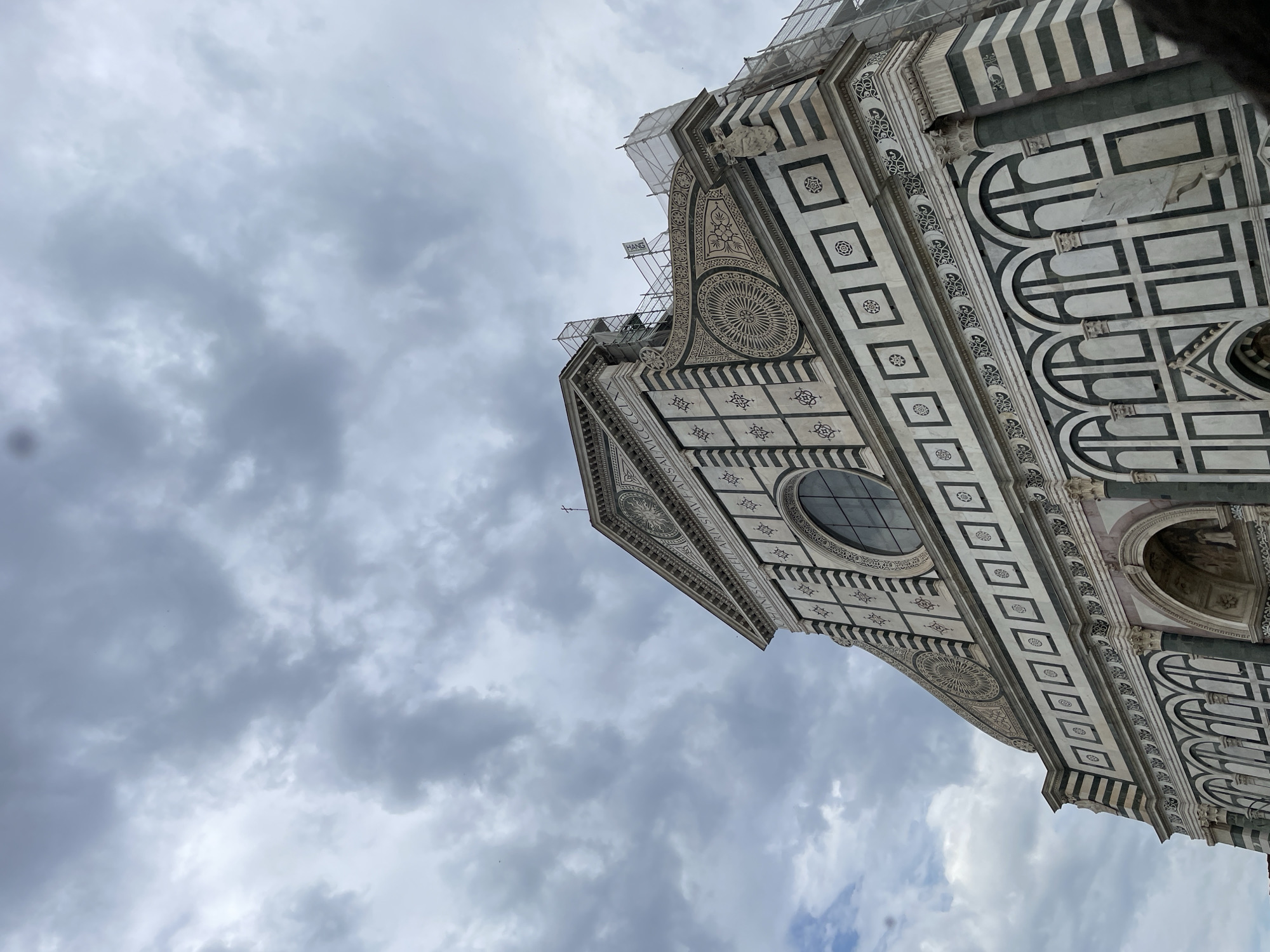 The image shows a low-angle view of the Basilica di Santa Maria Novella in Florence, Italy. The basilica's facade is made of white and green marble, with geometric patterns and decorative arches. The sky is overcast with grey clouds. Some scaffolding is visible near the top of the basilica, indicating ongoing maintenance or restoration work. There are no people visible in the image.