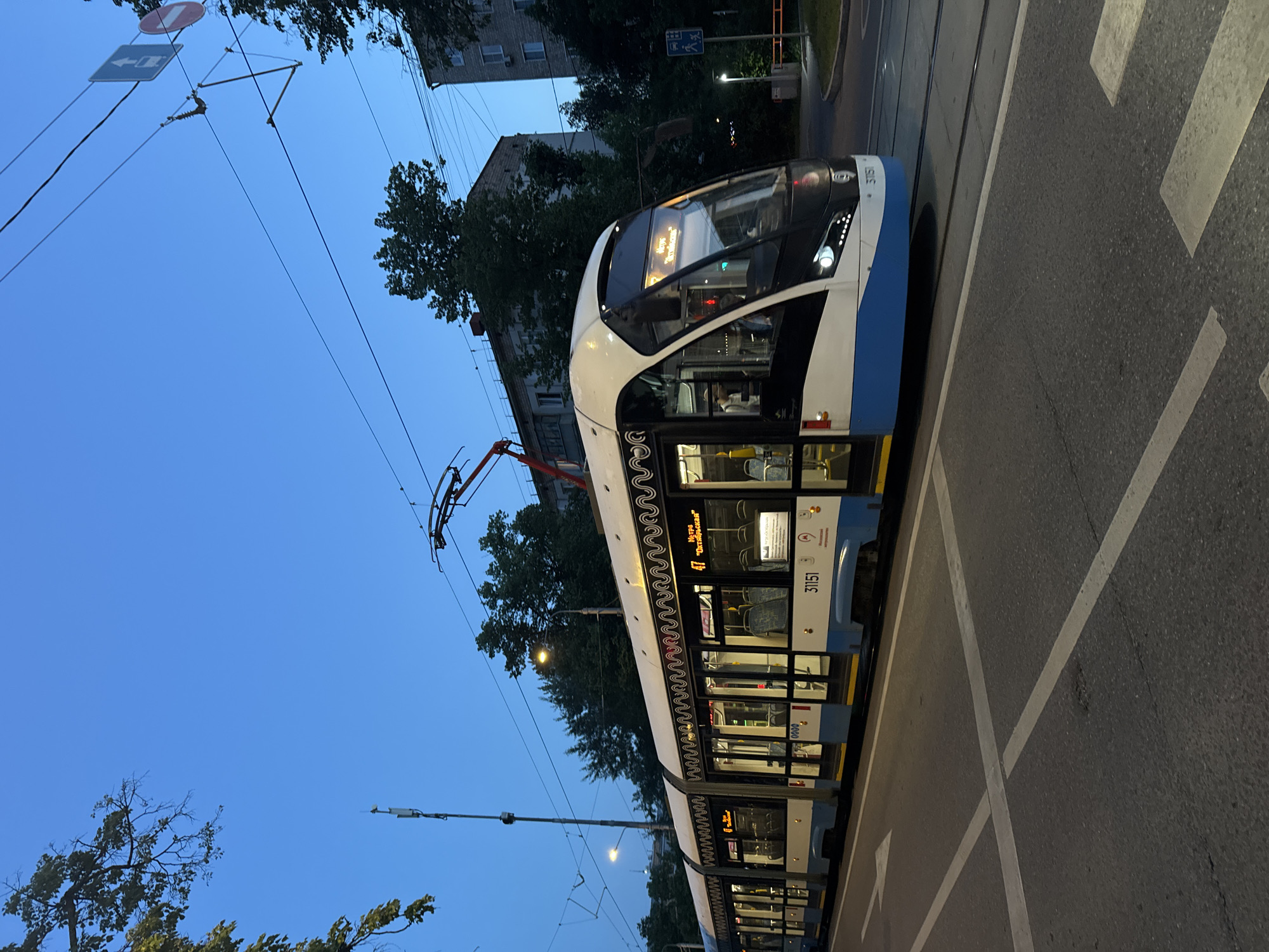 A white and blue tram with yellow accents is traveling on a street in Moscow, Russia, during the blue hour. The tram is modern, with large windows and illuminated interior lights. It has the number "31151" visible on its side, and a route display showing "47 метро 'Октябрьская'". The tram's pantograph is extended and connected to overhead power lines. The street is paved with asphalt and marked with white road lines, including a crosswalk. In the background, there are trees, buildings, a lamppost, and traffic signs. The sky is a deep blue, indicating dusk or early morning. The overall scene depicts urban public transportation in Moscow at twilight.