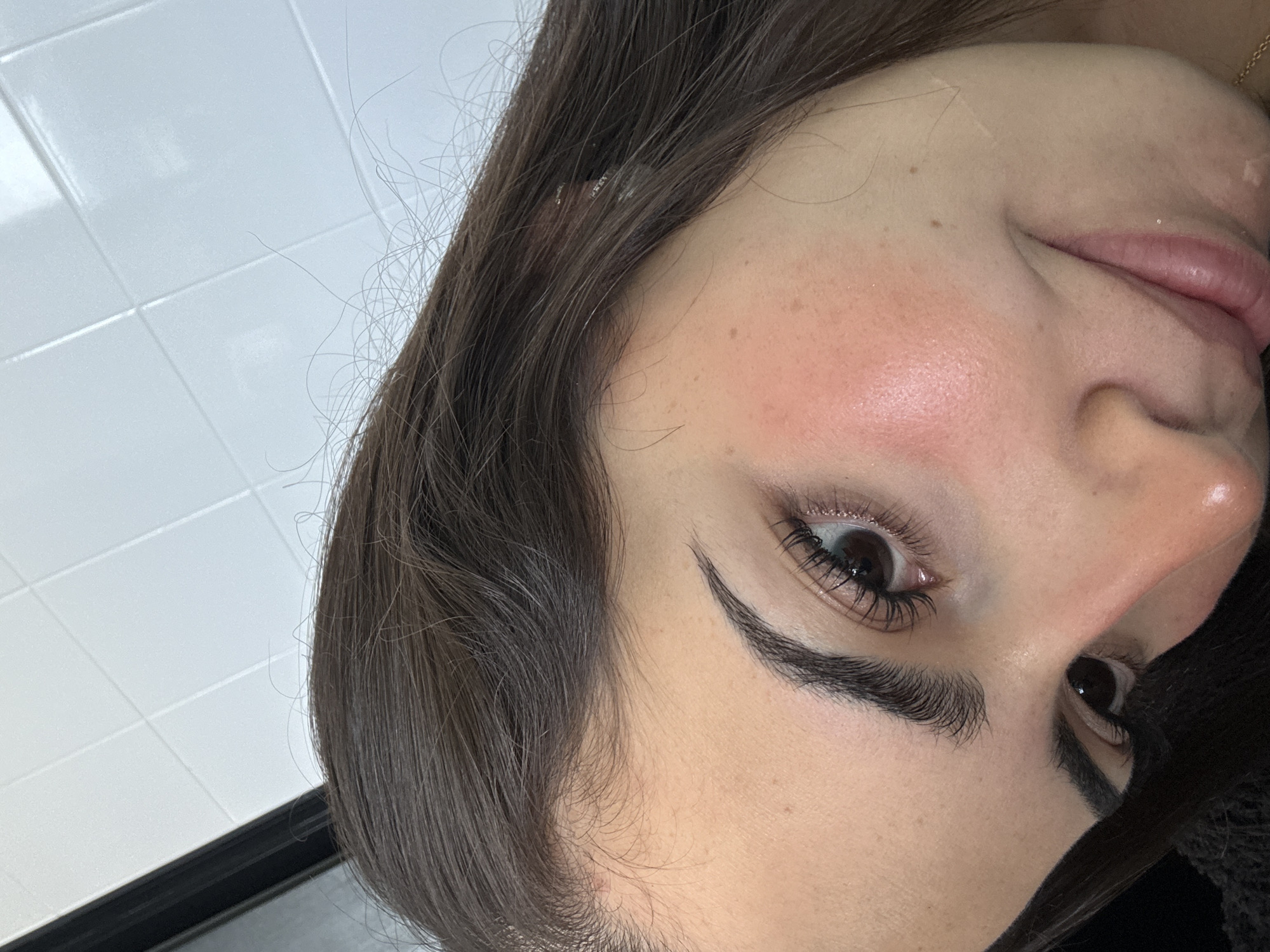 A close-up portrait shows a section of a person's face, tilted sideways at an approximate 90-degree angle. The subject is a woman with dark brown hair, several strands of which are loose around her face. Her left eye is prominently featured, with well-defined dark eyebrows and long, dark eyelashes suggesting mascara or extensions. A subtle shimmer or glitter is visible on the eyelid near the outer corner. Her cheek displays a flush of pink blush, and the skin has some visible freckles and moles. Part of her nose and upper lip are also seen, with the lips appearing a natural pink. A hint of a gold chain is visible near the bottom right edge of the frame. The background consists of a bright white tiled surface, indicating an indoor setting, possibly a bathroom or a similar tiled space. The overall impression is a posed shot, focusing on facial features and makeup. No specific activity beyond posing is discernible. The image was captured in San Bernardino Tlaxcalancingo, Mexico. There is no visible text.
