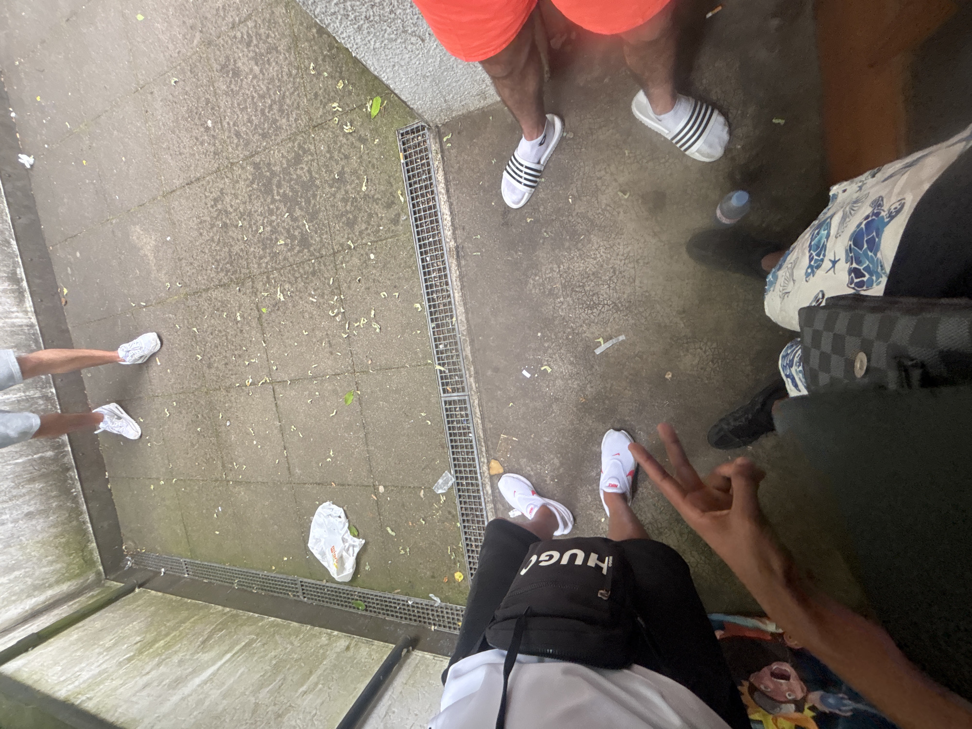 The image depicts a group of people standing on a paved surface with a drainage grate in Liestal, Switzerland. There are at least three people visible. One individual wears orange shorts and white Adidas slide sandals with black stripes, with socks. Another person, wearing dark clothing and white Nike shoes with red accents, is making a hand gesture. A third person is partly visible, wearing dark clothing and a white and blue patterned bag. There is a trash bag and loose debris scattered on the ground. The environment appears to be an urban outdoor area.
