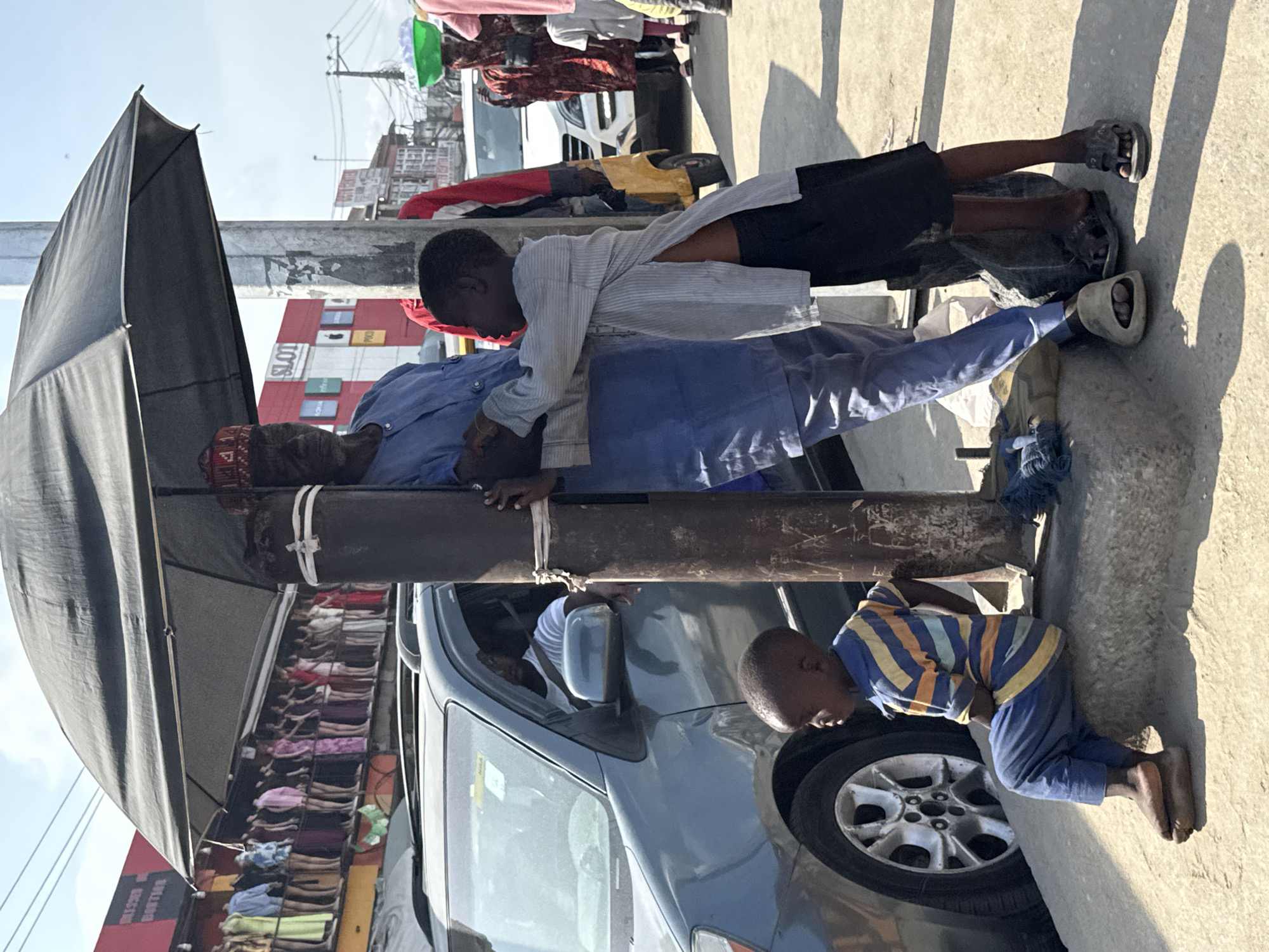 A scene captured in Lagos, Nigeria, showcases an urban street setting with a variety of activities. An adult man, with a red fez-type hat, wearing a light blue shirt and blue pants, is lying on a metal pole in the foreground. The pole is held up by a black umbrella. A second, younger male is resting on top of the older man. He is wearing a light-colored striped shirt and black shorts, and flip-flops. Another child, in a blue and yellow striped shirt and blue pants, kneels next to the front passenger tire of a gray car. The gray car obscures the street in the foreground. Clothes, shoes, and accessories displayed on a makeshift rack are visible in the background. A building with a red and white facade is noticeable further behind.
