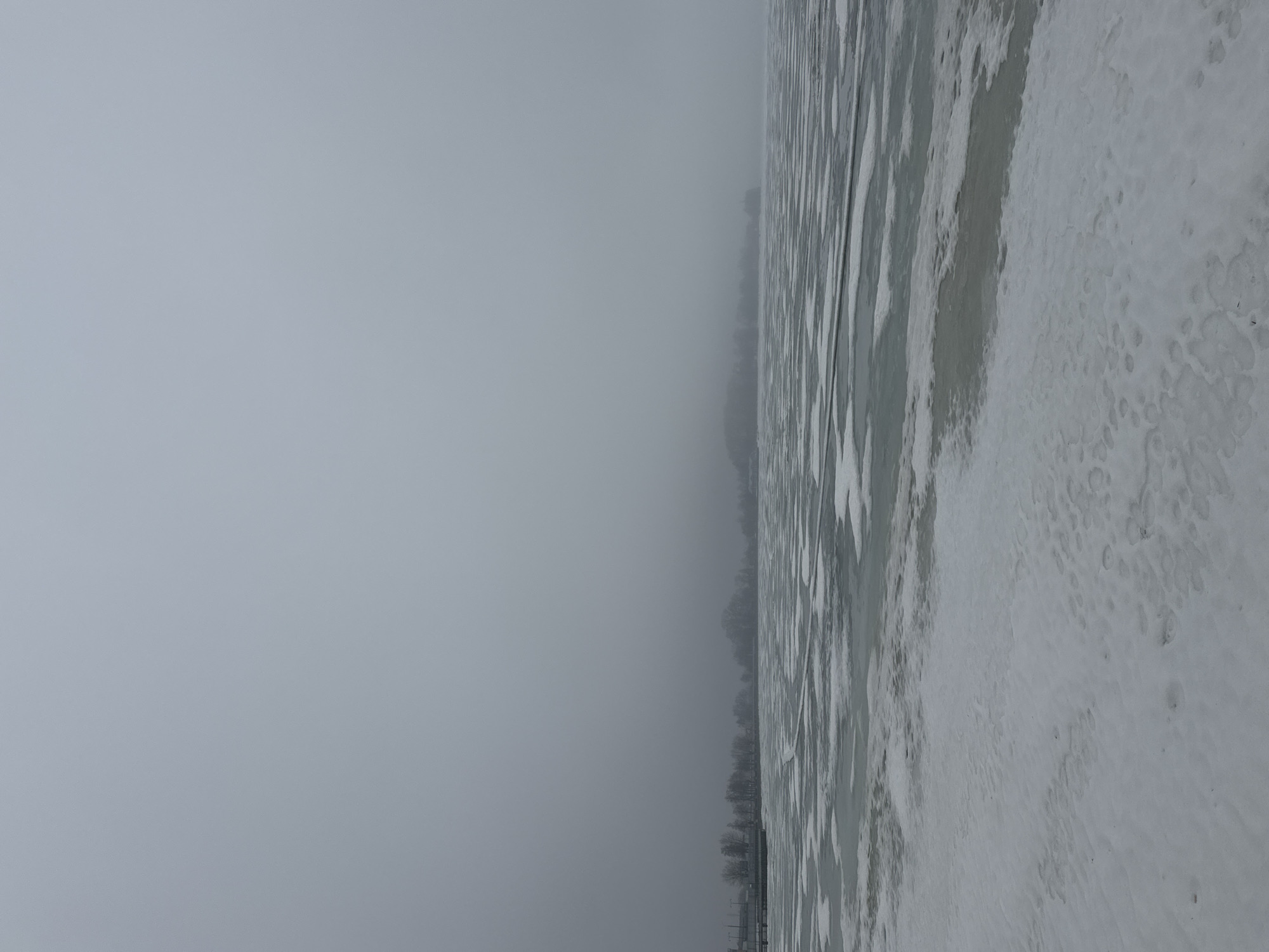The image depicts a snow-covered, partially frozen body of water, possibly a lake or wide river, under a heavy, overcast sky. In the middle ground, the water transitions into a densely packed ice surface covered with a layer of snow. There are multiple visible footprints on the snow-covered ice. Along the distant shoreline, a blurred treeline and a few buildings can be faintly discerned. The overall environment suggests a cold, winter day. The scene suggests the city of Votkinsk, Russia, given the prompt’s instructions, but without corroborating information within the image.