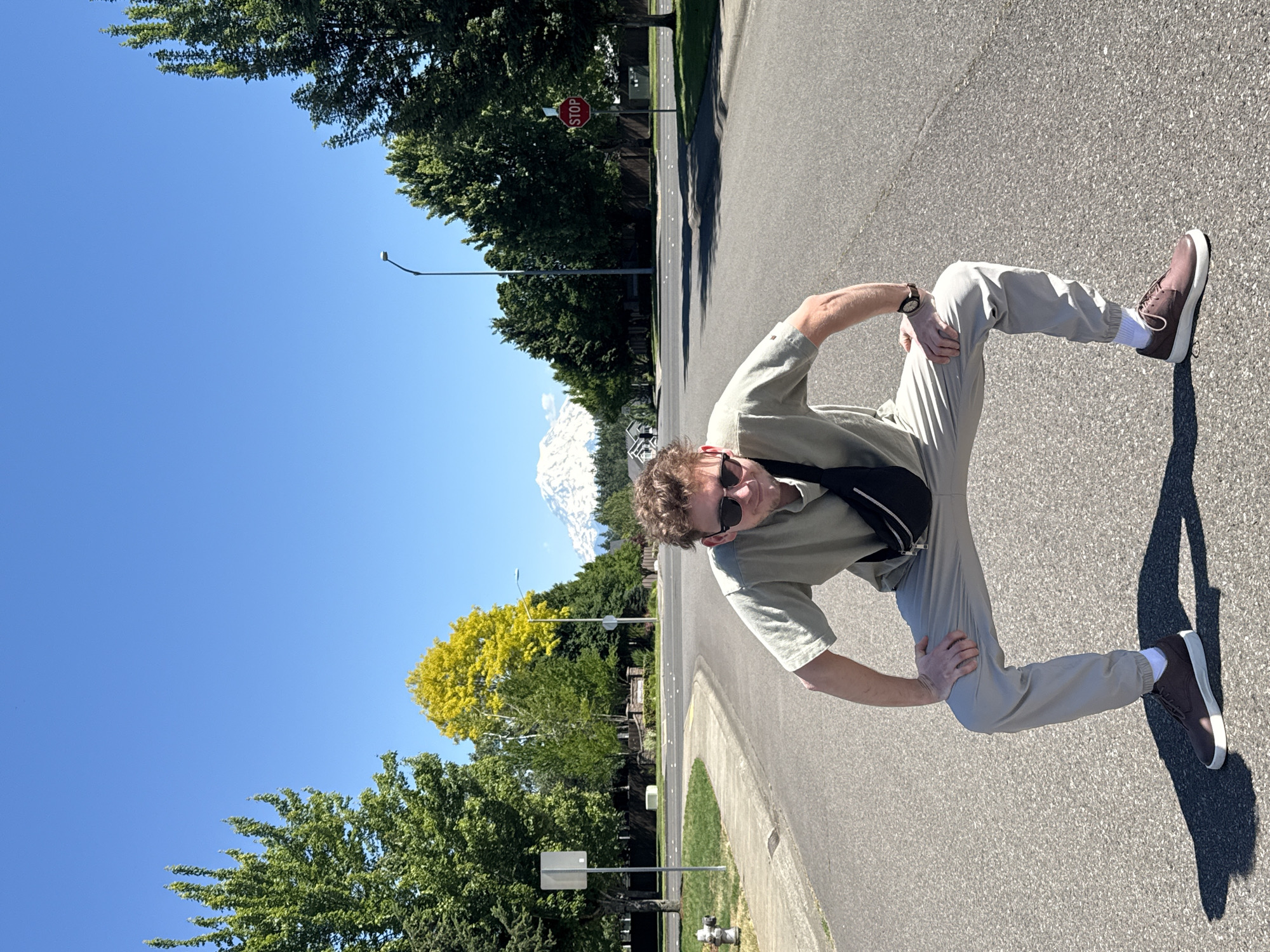 A man with curly brown hair and sunglasses crouches in the middle of a residential street in South Hill, Washington, United States of America. He wears a light green t-shirt, light gray pants, and brown shoes with white soles. A black fanny pack is slung across his chest. His right hand rests on his right knee, and his left arm is bent at the elbow. The street is paved and lined with trees on both sides. In the background, a snow-capped mountain, likely Mount Rainier, is visible under a clear blue sky. A stop sign is posted on the right side of the street, and a fire hydrant is on the left. The sun casts long shadows on the asphalt.