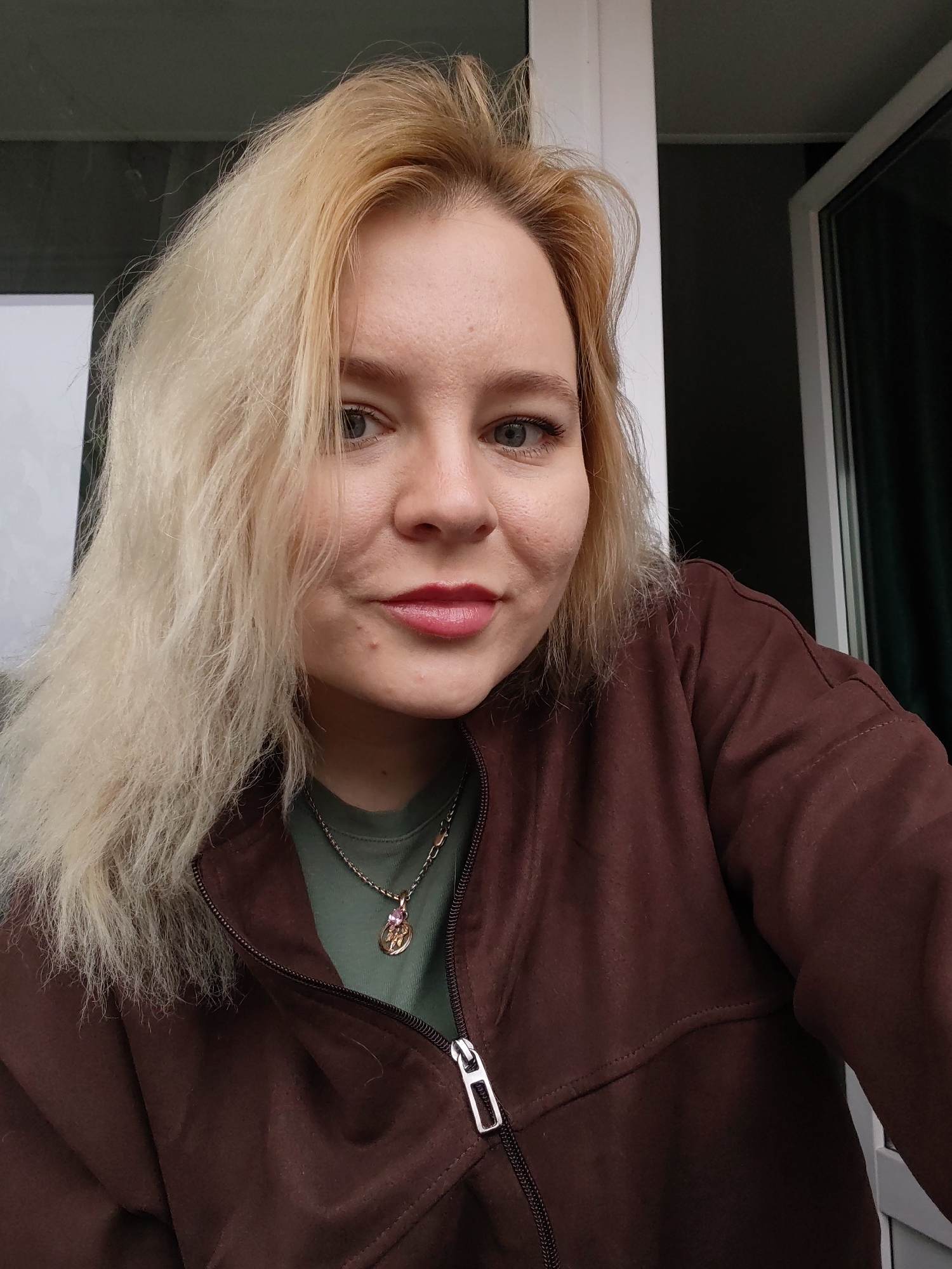 A blonde-haired woman is in the foreground of the shot, wearing a brown jacket and green t-shirt. A necklace with a pink stone is visible. Behind her are a white wall and window/door. The location in the city of Izhevsk, Russia is not identifiable based on the visual information provided.
