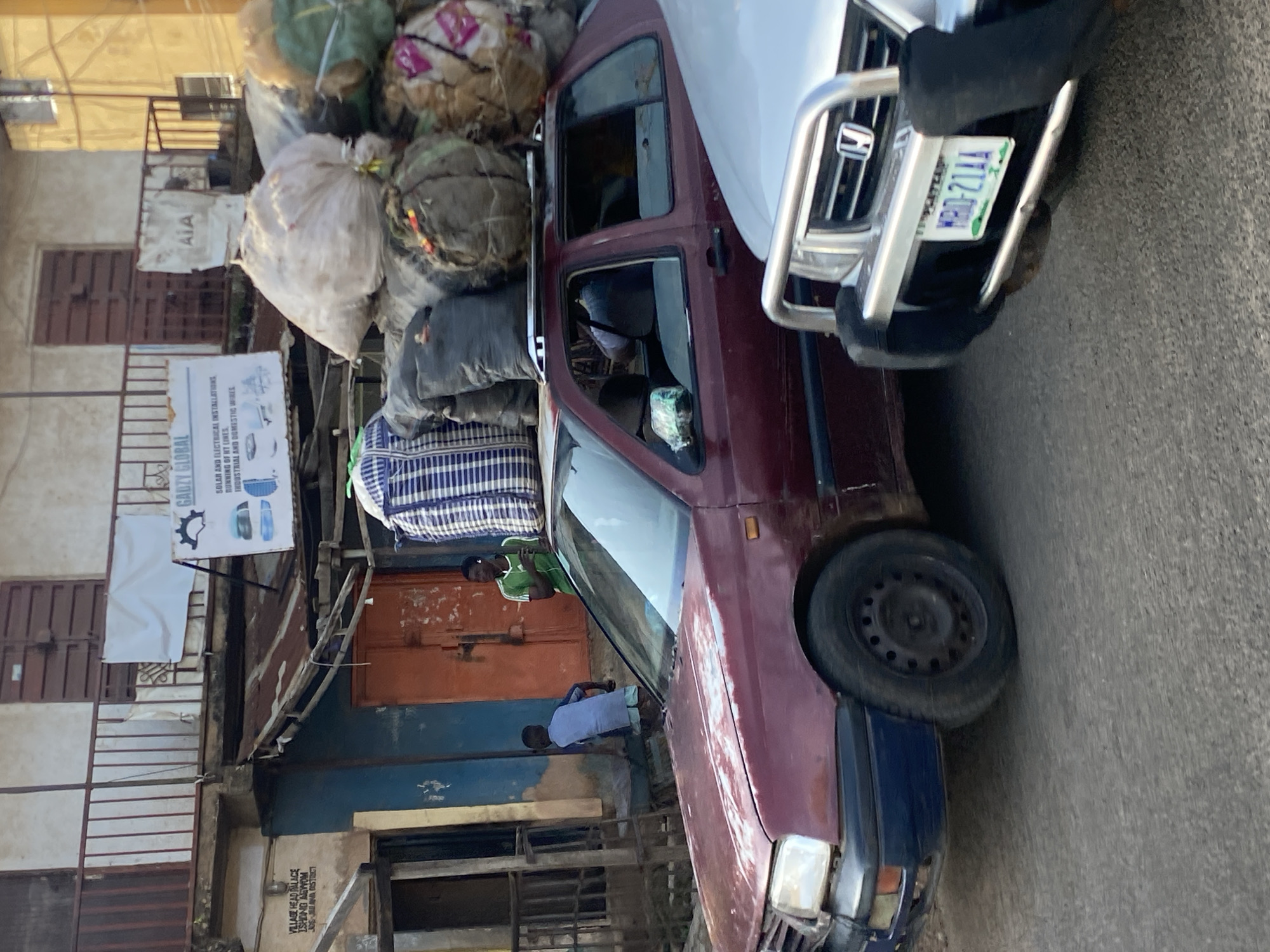 In Gwong, Nigeria, a maroon sedan is heavily loaded with various items on its roof. These include large, cylindrical bags wrapped in netting, as well as a large, checkered blue and white duffel bag. Two young boys are near the car, one wearing a light blue shirt and shorts, the other a green t-shirt and shorts. A third person, possibly a man, is also visible in the distance. The car is parked on a paved road in front of a building with a balcony and an orange door. A silver SUV is parked in front of the sedan. The building's facade is weathered and shows signs of age, with peeling paint and a rusted corrugated metal roof section. A sign on the building reads "GADZY GLOBAL".
