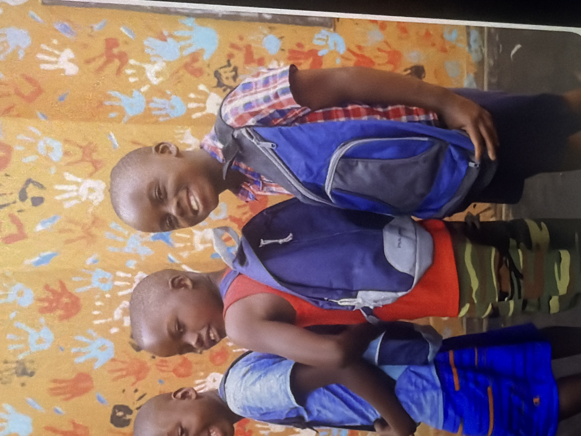 Three young Black boys are standing in front of a wall painted with colorful handprints in an outdoor setting. The boy on the left is wearing a blue t-shirt and blue shorts with orange stripes. The middle boy is wearing a red tank top and camouflage pants, and he has a blue backpack on. The boy on the right is wearing a red and blue plaid shirt and a blue and gray backpack. All three boys are smiling. The background wall is a vibrant yellow with orange, blue, and white handprints scattered across it. The ground appears to be a dark, textured surface, possibly asphalt or concrete. The lighting suggests it is daytime, with soft shadows indicating diffused sunlight.