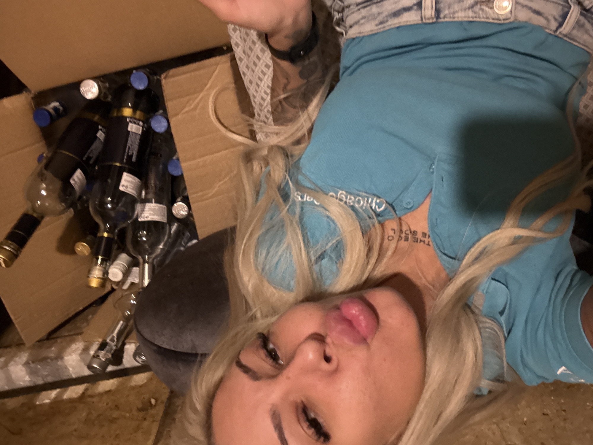 A photograph depicts a woman with blonde hair, wearing a blue polo shirt with white lettering "Chicago Bears" near the collar, and a denim bottom. She is positioned beside a cardboard box filled with several wine bottles. The location or environment cannot be determined as the photo does not depict any landmarks or clues to suggest a specific city, including Plovdiv, Bulgaria.
